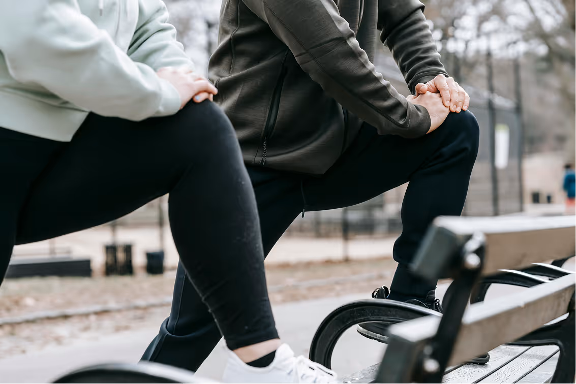 Two people stretching their legs outdoors with one foot resting on a bench.
