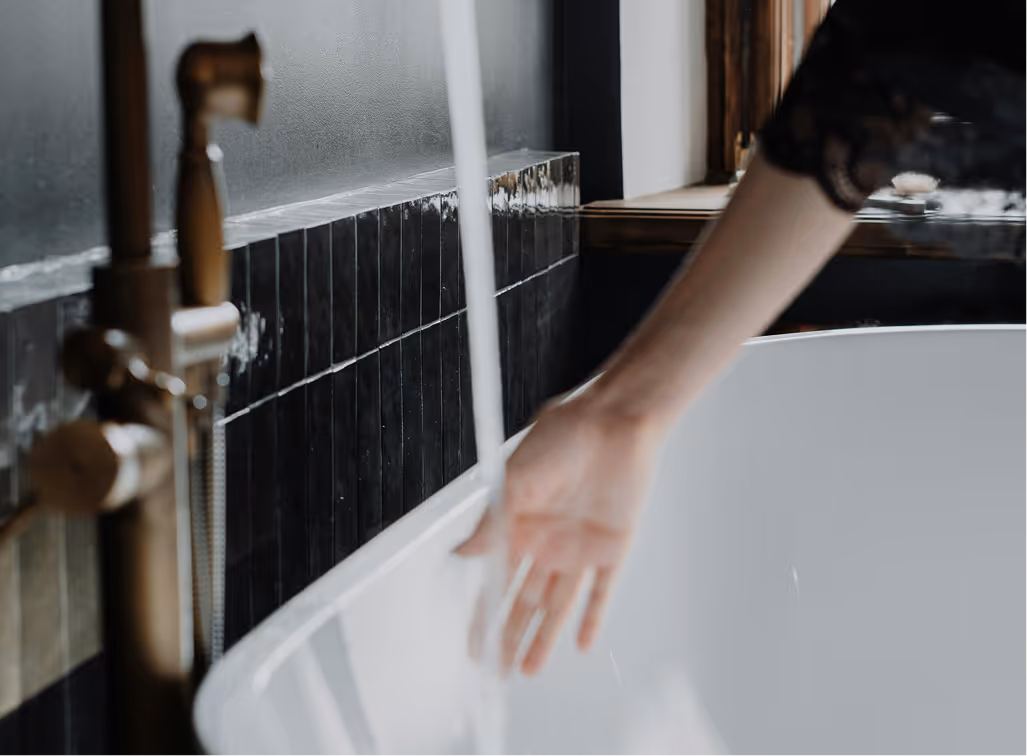Person's hand reaching under running water from a faucet into a white bathtub with black tiled wall in the background.