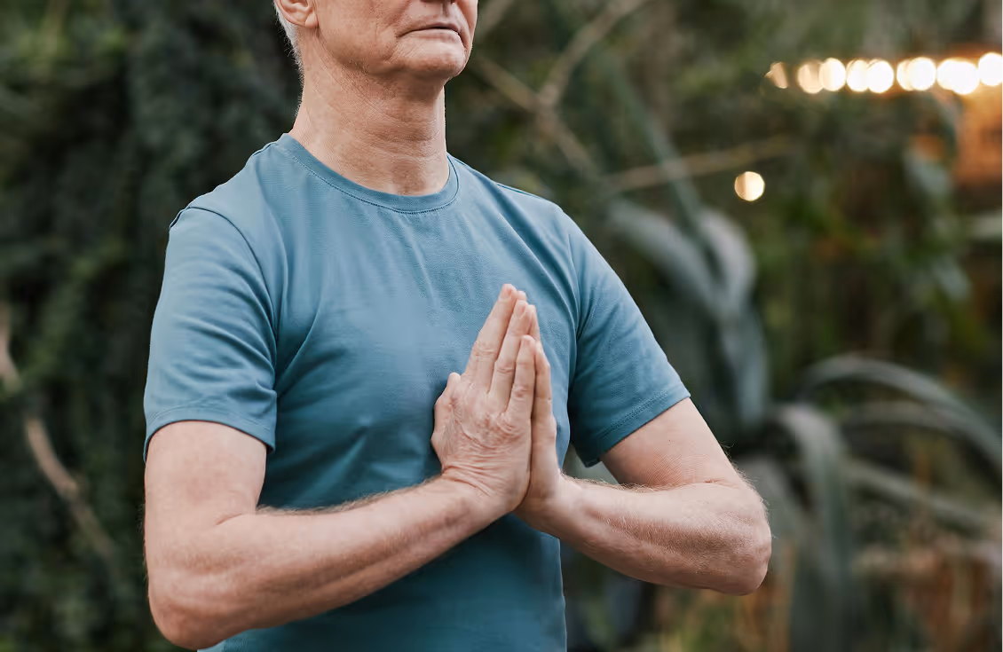 Middle-aged man in a blue t-shirt holding hands in a prayer or meditation gesture outdoors.