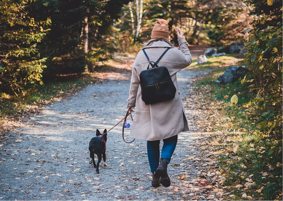 Person wearing a beige coat, brown hat, and black backpack walking a small black dog on a leash along a leafy forest path.