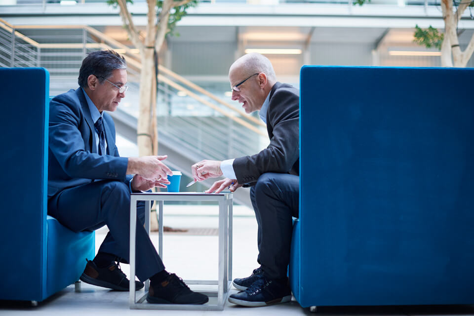 Two businessmen in suits sitting inside blue partitioned chairs across a small table in an office setting.