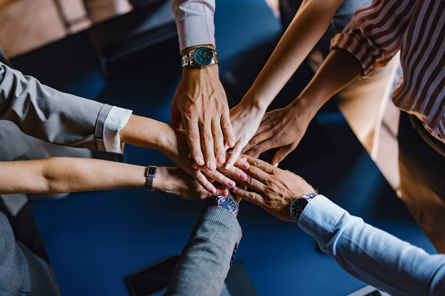 A top-down view of multiple diverse business professionals stacking their hands together in the center of a table to show teamwork and unity.
