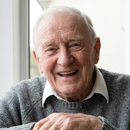 Portrait d'un homme âgé souriant avec un pull gris et une chemise blanche près d'une fenêtre.