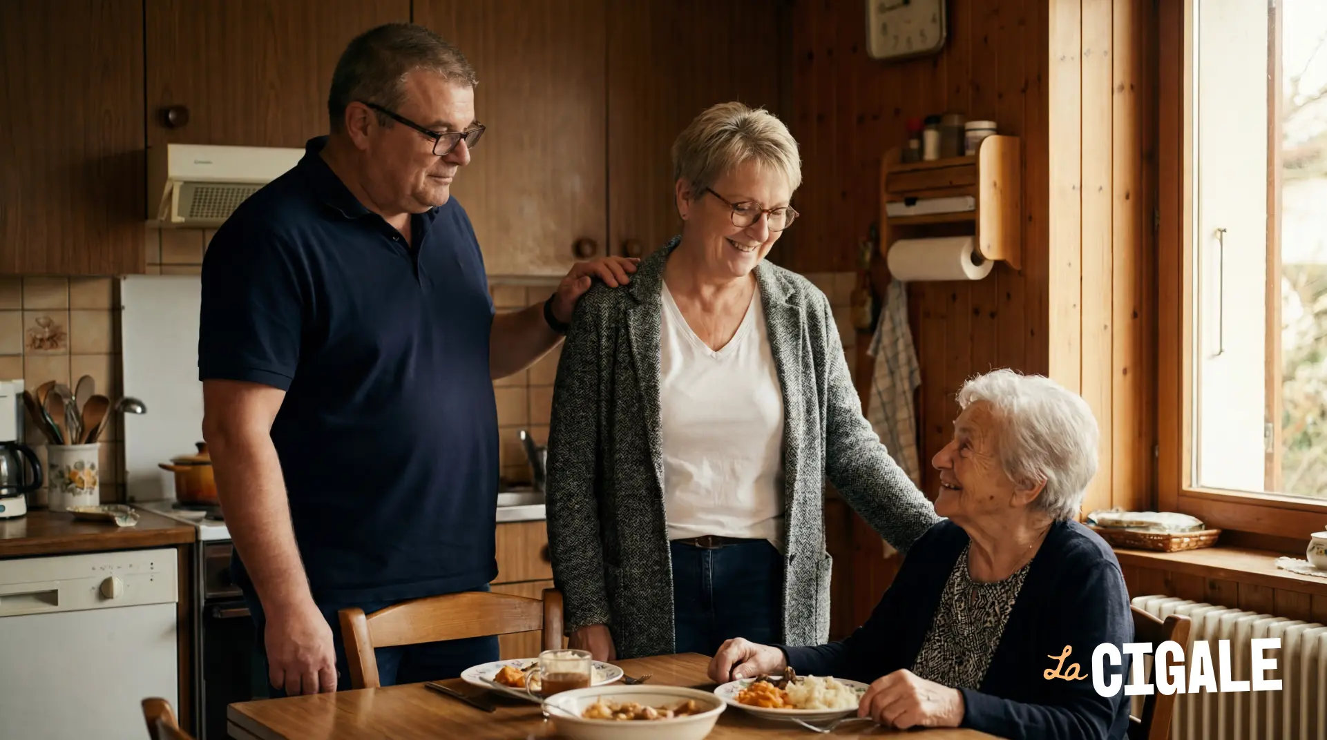 Un couple souriant soutient une femme âgée assise à table dans une cuisine chaleureuse.