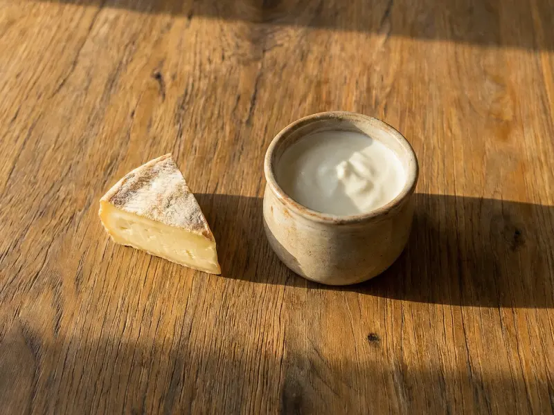 Un morceau de fromage à croûte blanche à côté d'un pot en céramique avec une crème blanche.