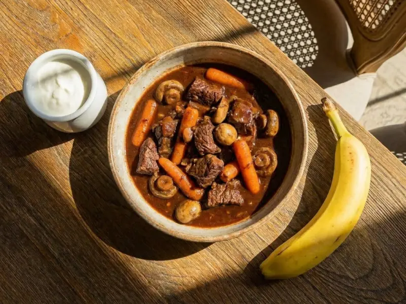 Plat de bœuf mijoté avec carottes, champignons, une banane à côté et un pot de sauce blanche.