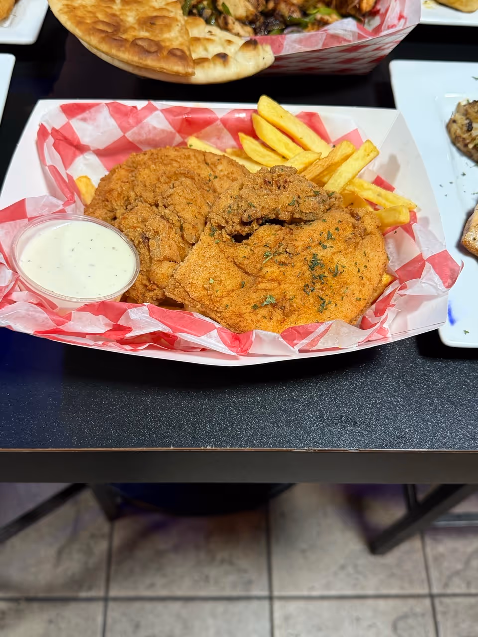 Fried chicken tenders with French fries and ranch dipping sauce in a red-checkered paper food tray.