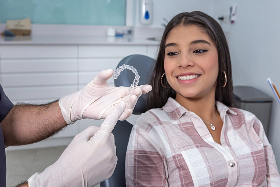 Woman smiling in dentist chair as dentist holds a clear dental aligner with gloved hands.