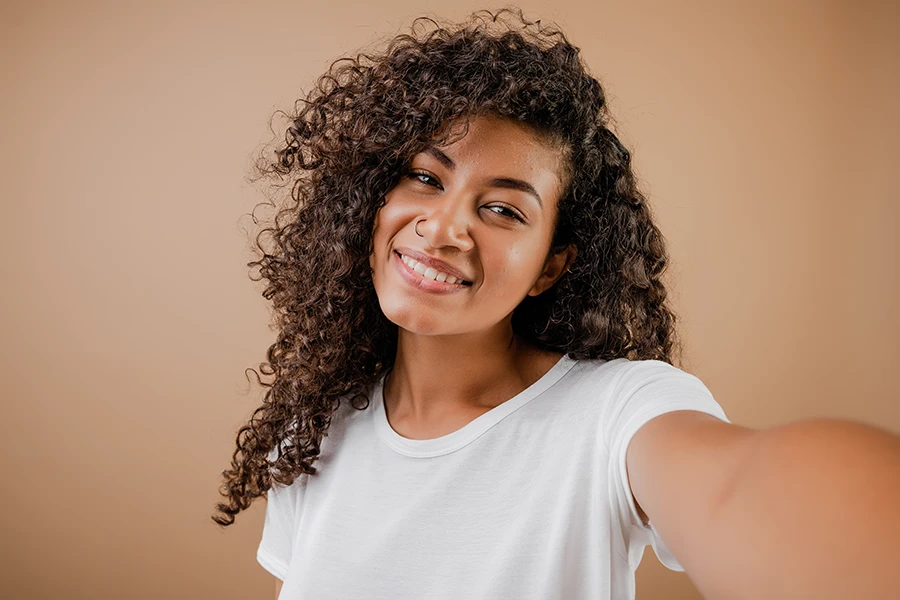 Smiling woman with curly hair taking a selfie against a beige background.
