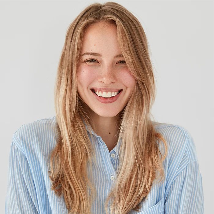 Smiling young woman with long blonde hair wearing a blue striped shirt against a plain background.