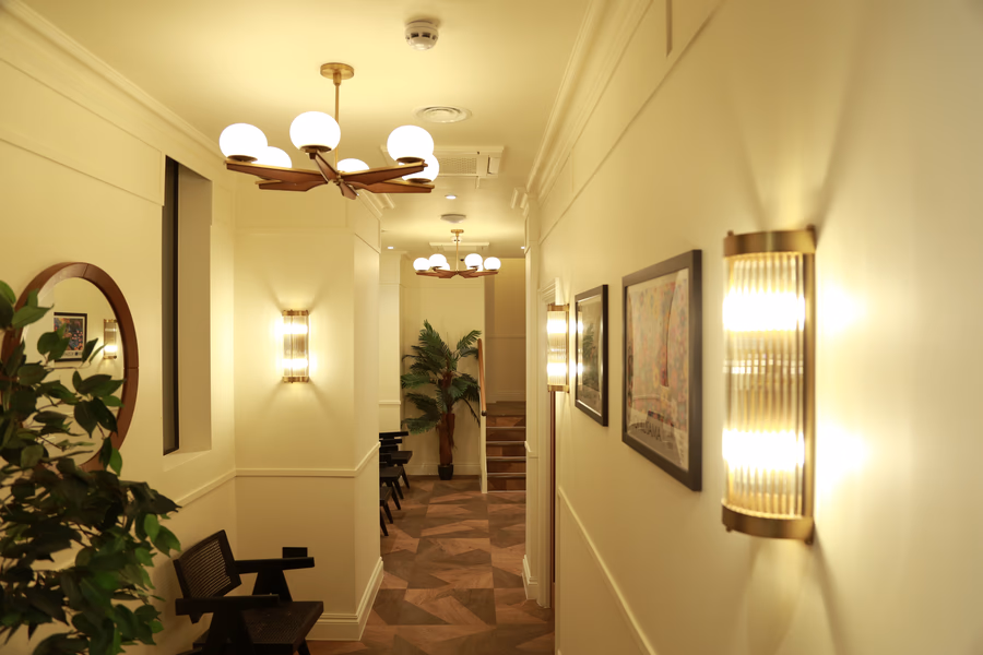 Well-lit modern hallway with wooden flooring, wall sconces, round mirror, framed art, and potted plants.