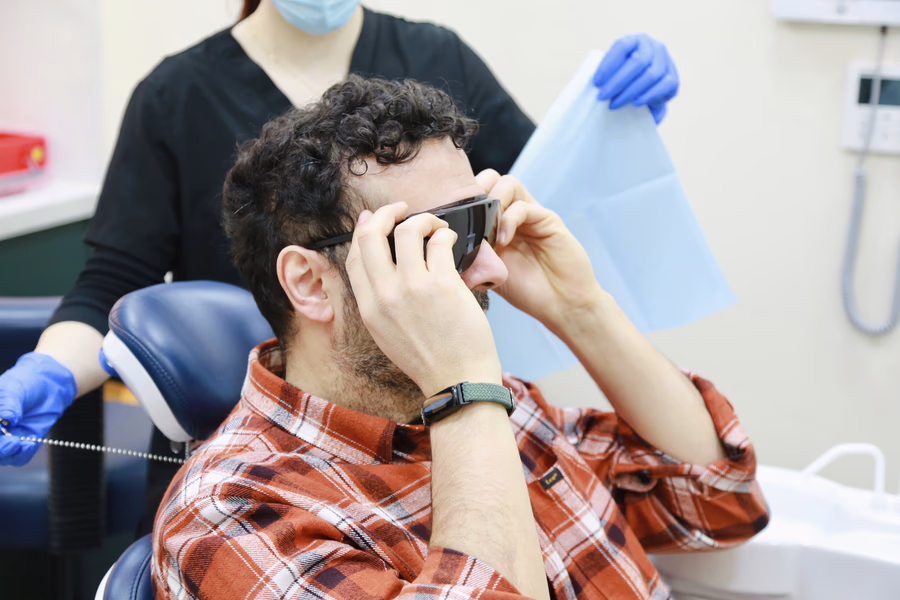 Man wearing a red plaid shirt and protective glasses sitting in a dental chair with a dentist behind him.