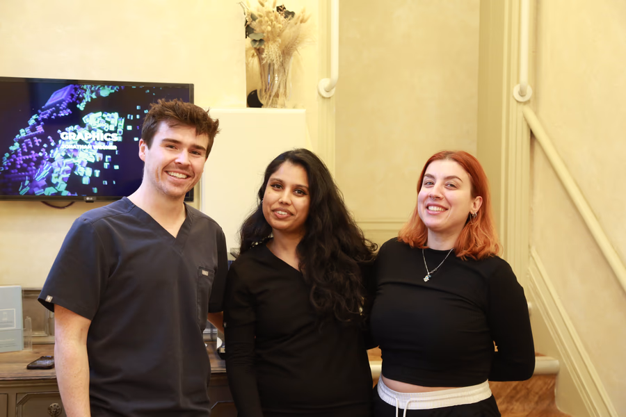 Smiling man in scrubs and two women in black tops standing indoors in front of a TV and vase with dried flowers.
