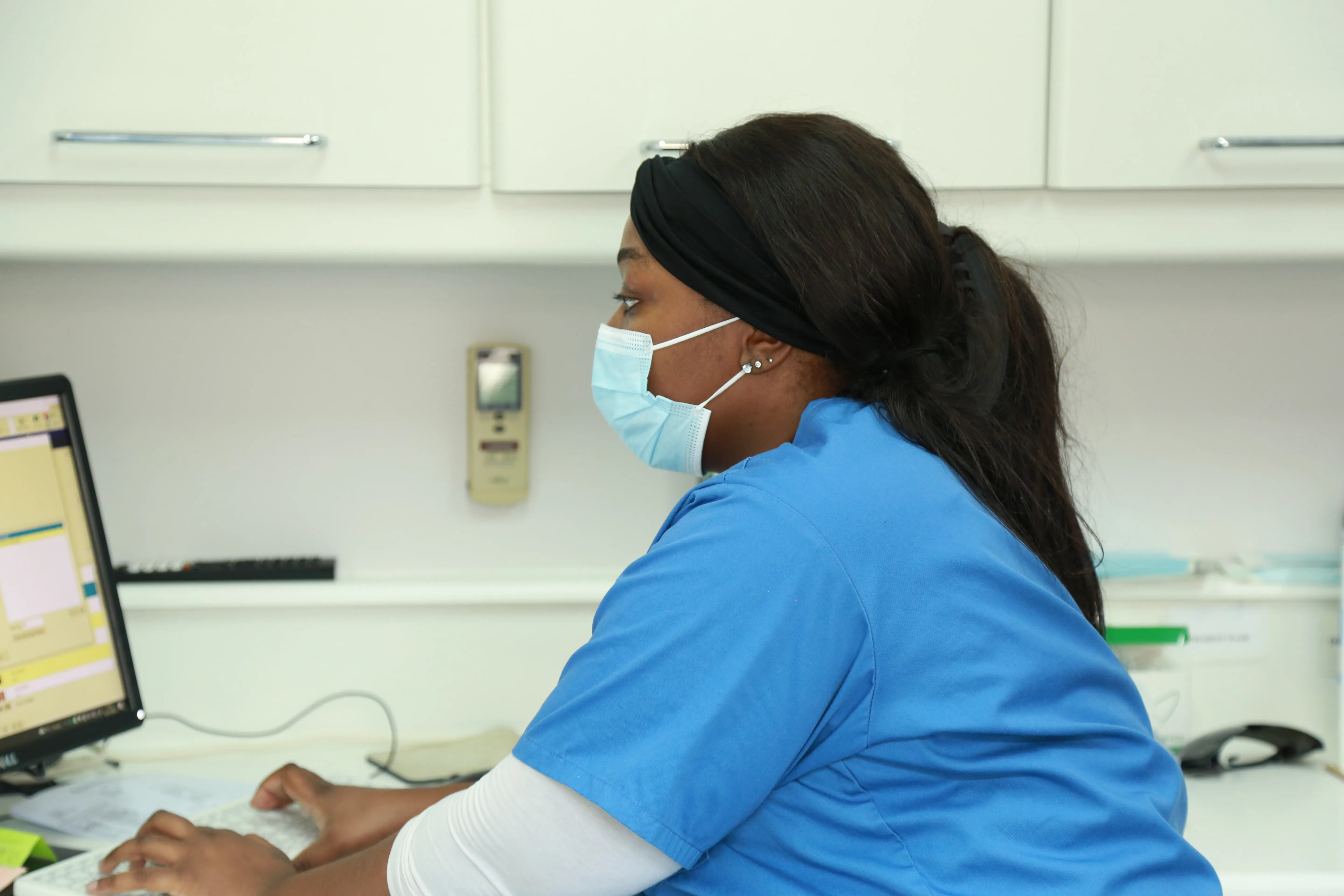 Healthcare worker in blue scrubs and face mask typing on a computer keyboard.