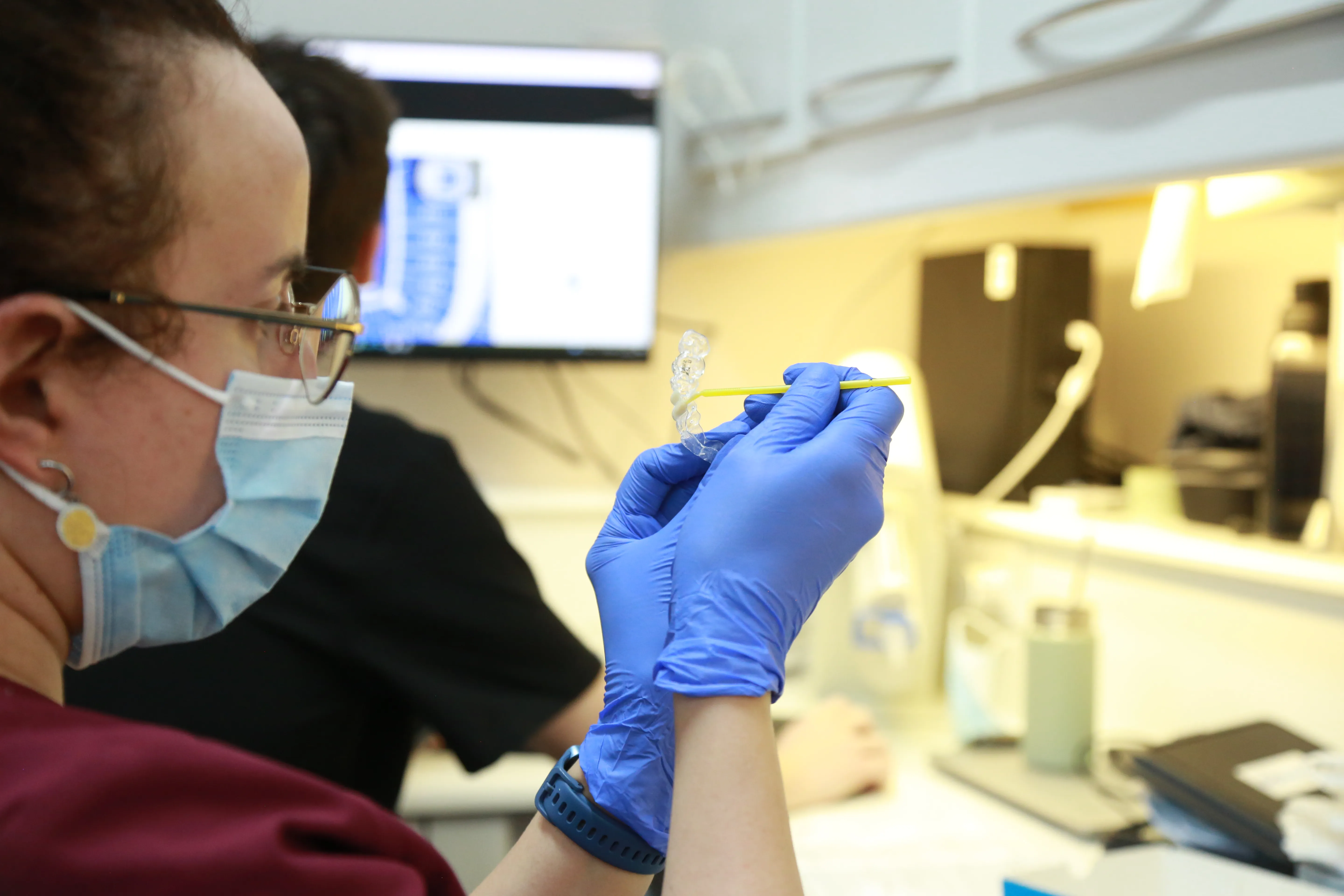 Dental professional wearing gloves and mask holds a clear dental aligner and yellow tool in a clinic.