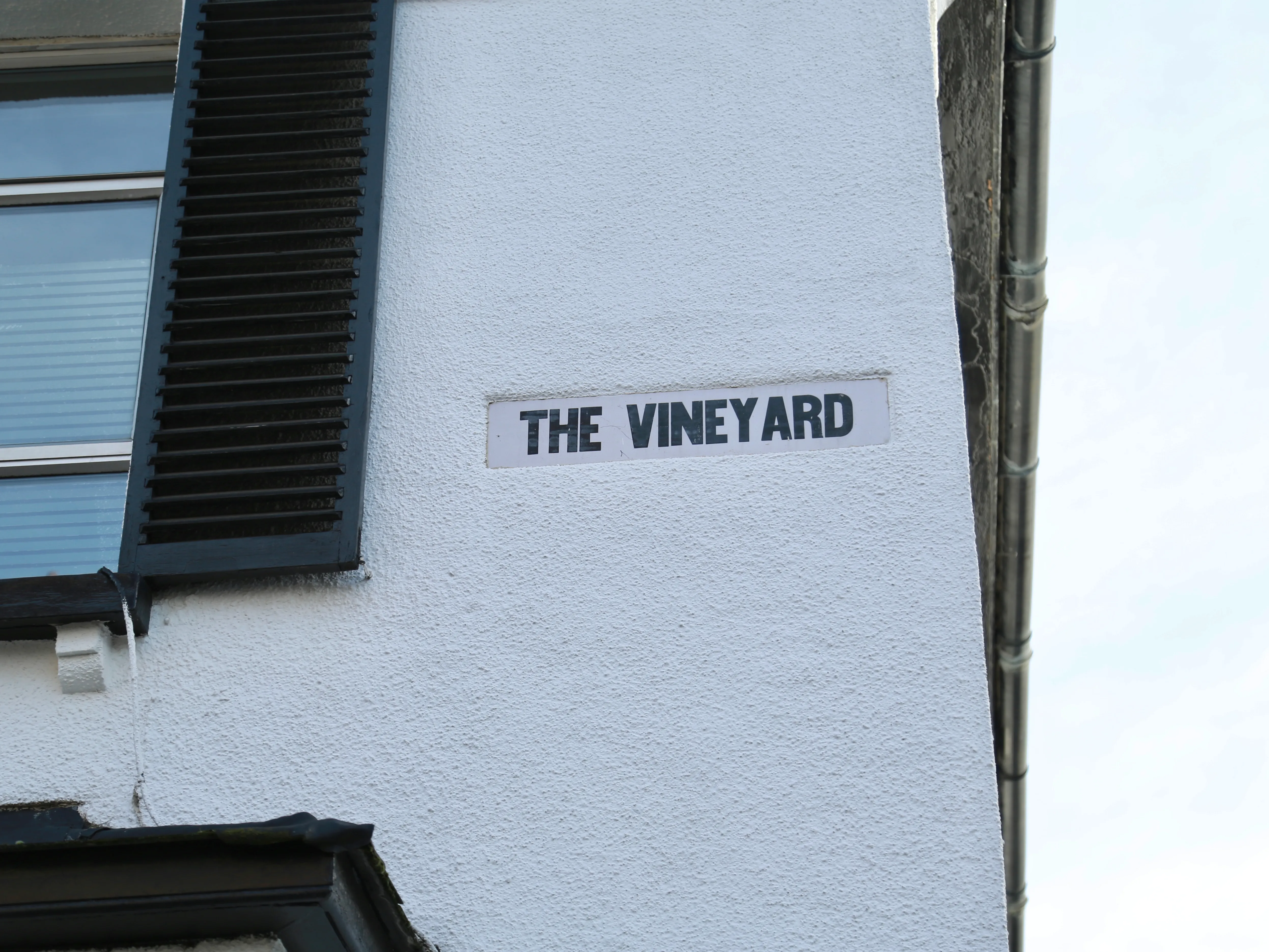 White building wall with a sign reading 'THE VINEYARD' next to a window with black shutters.