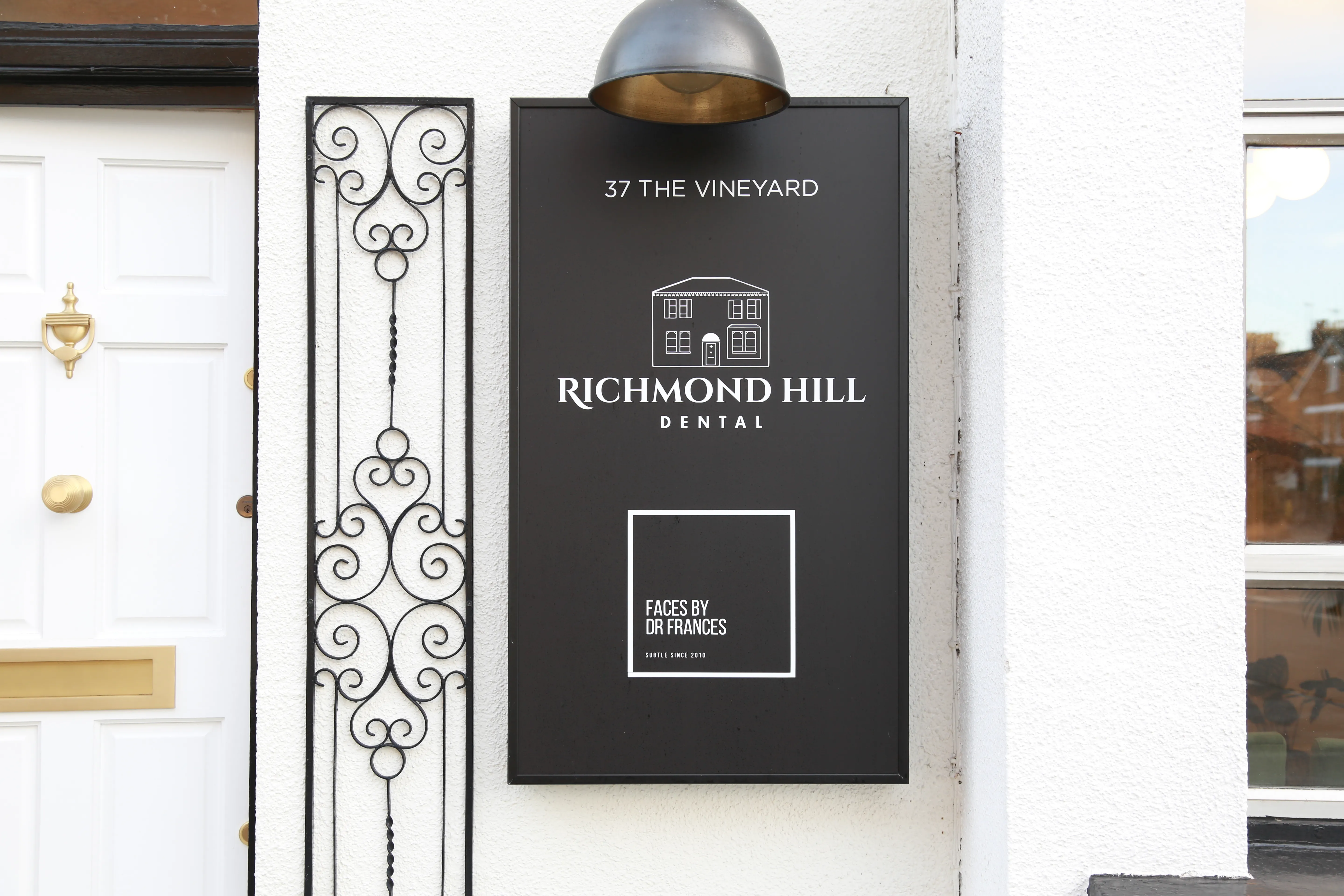 Black sign on white wall for Richmond Hill Dental at 37 The Vineyard with door and window nearby.