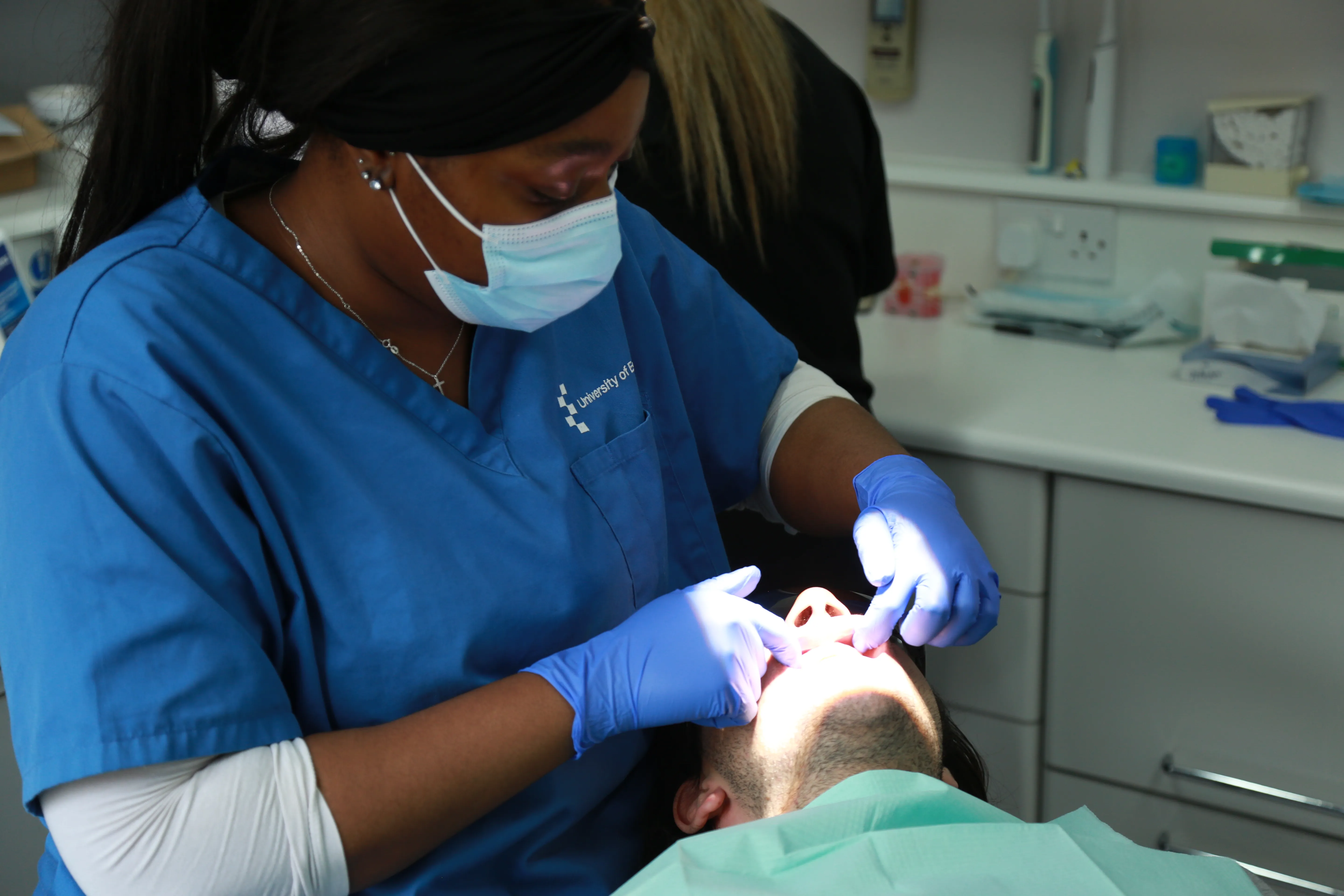 Dentist wearing blue scrubs and mask examining a patient's mouth in a dental clinic.