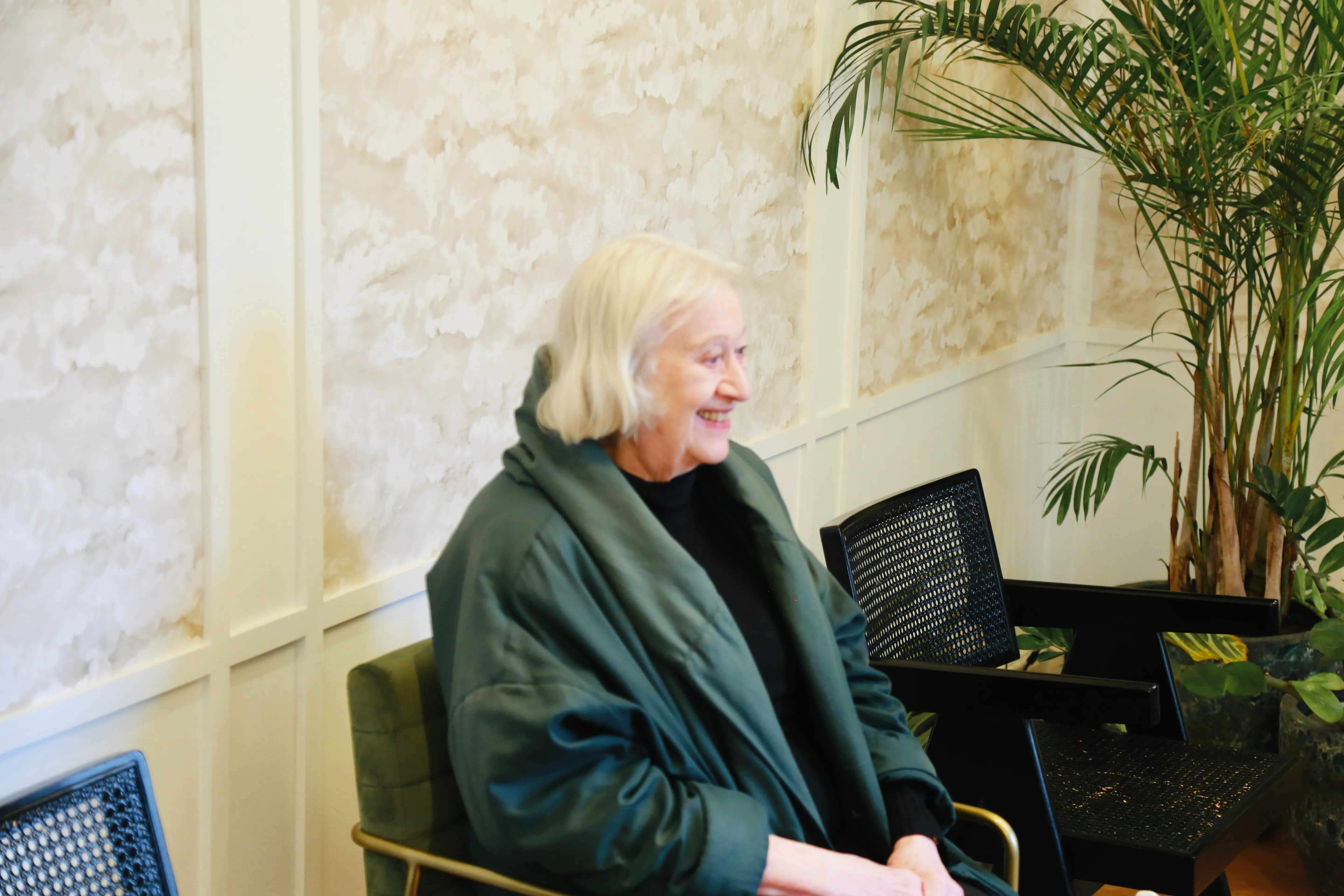 Smiling elderly woman with white hair in dark green coat sitting on a chair indoors near plants.