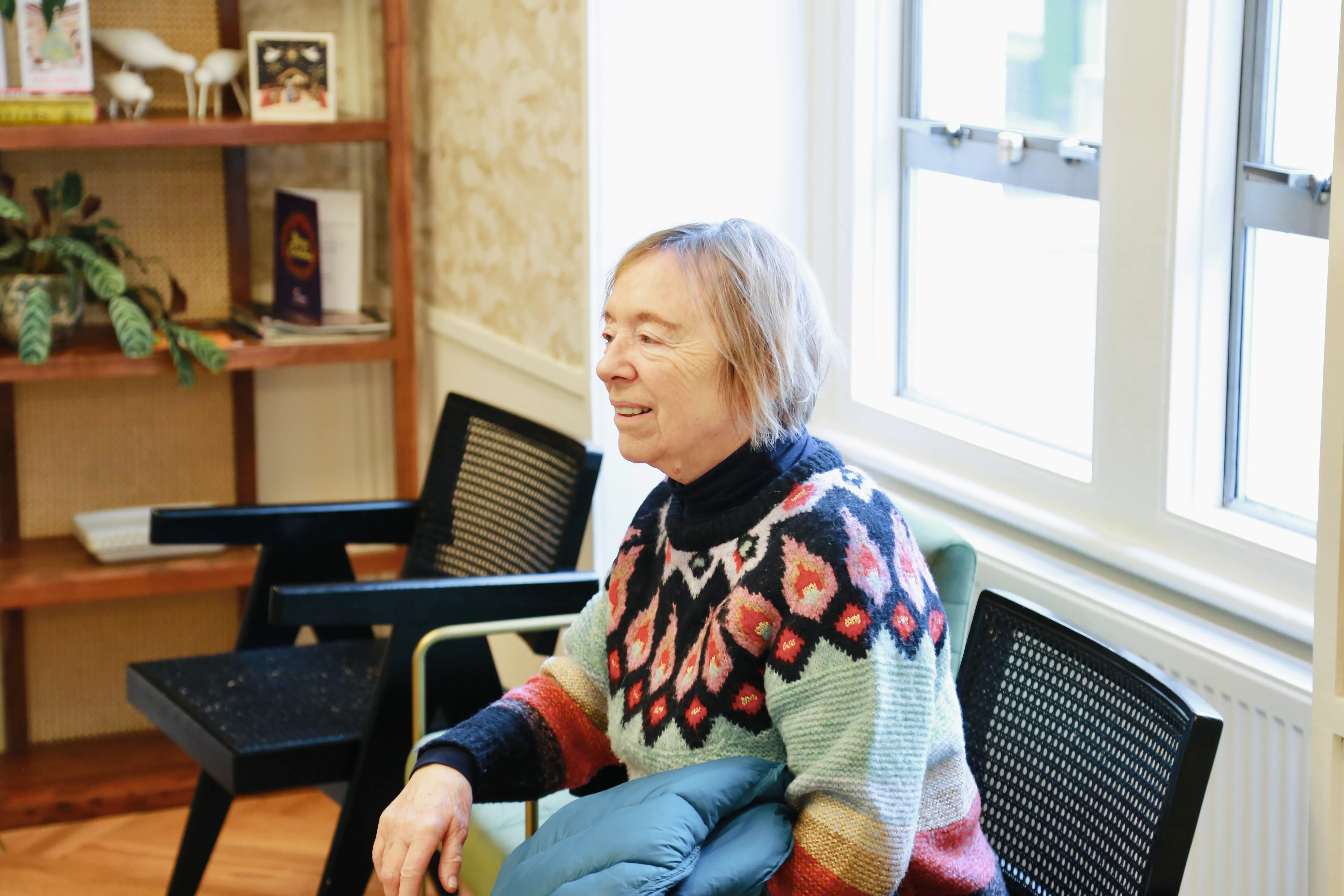 Smiling elderly woman wearing a colorful sweater sitting indoors near windows and holding a jacket.