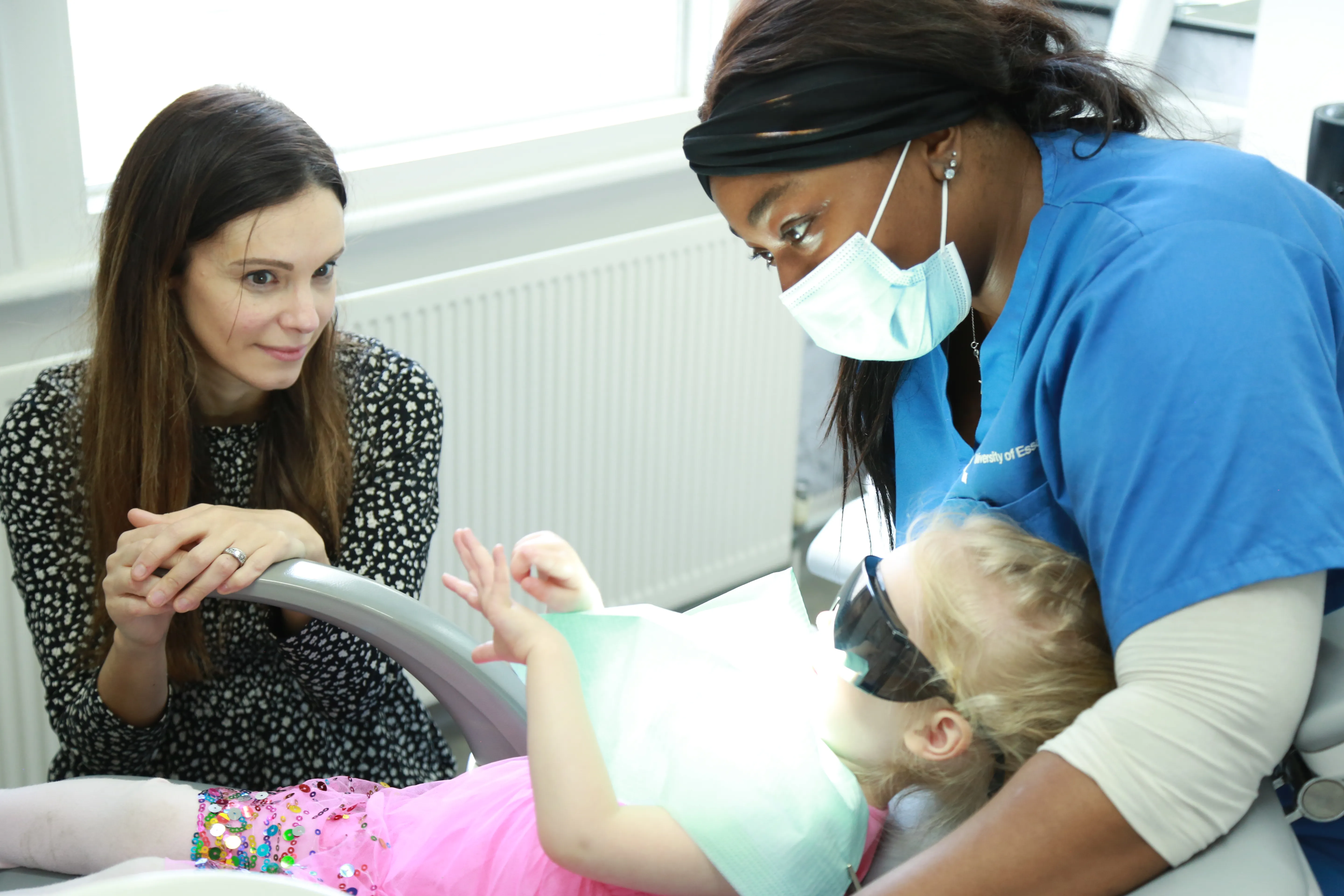 Dentist in blue scrubs wearing a mask examining a young girl wearing protective goggles and pink clothes.