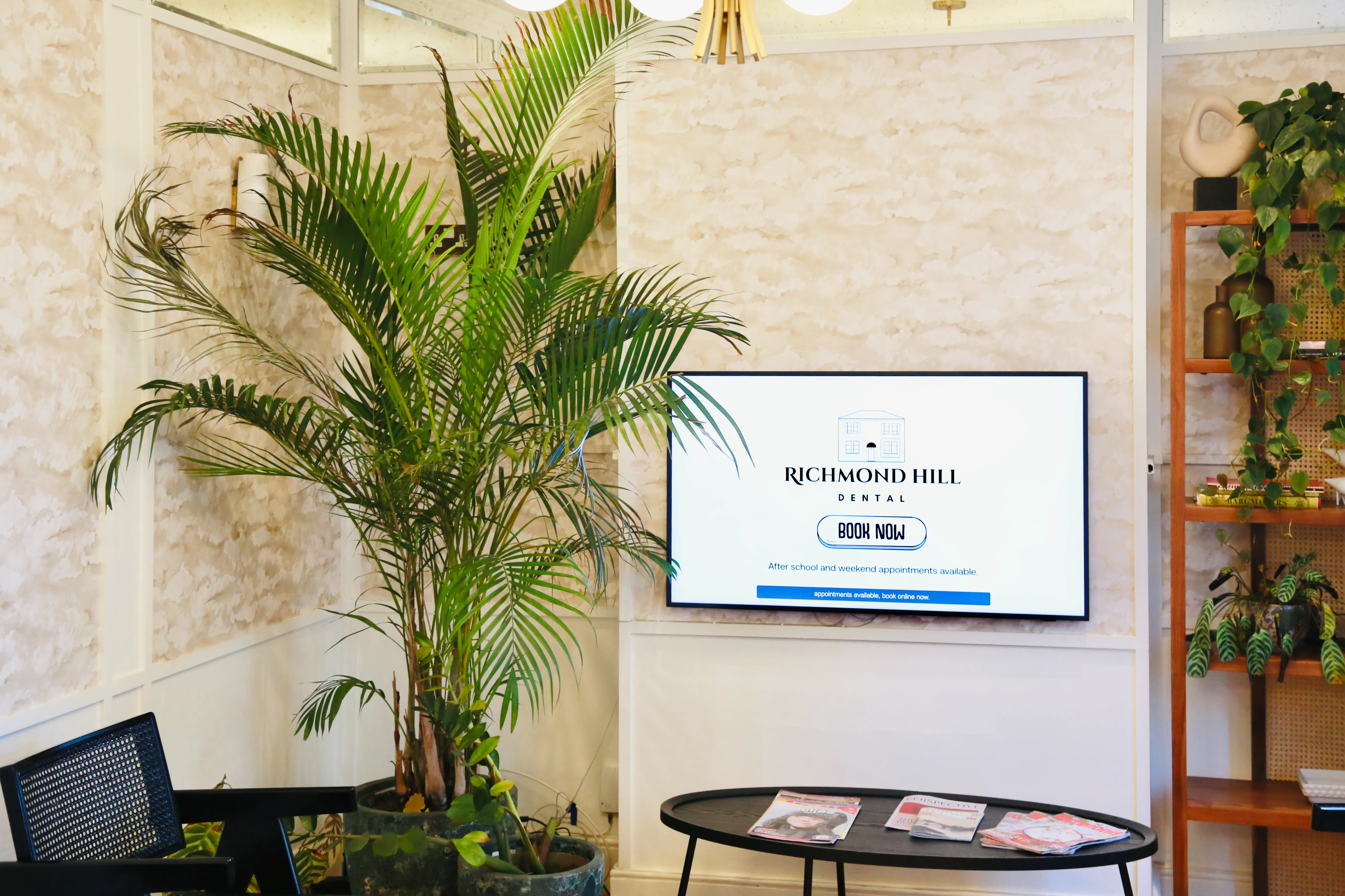 Waiting room with potted plants, a TV displaying Richmond Hill Dental and a black table with magazines.