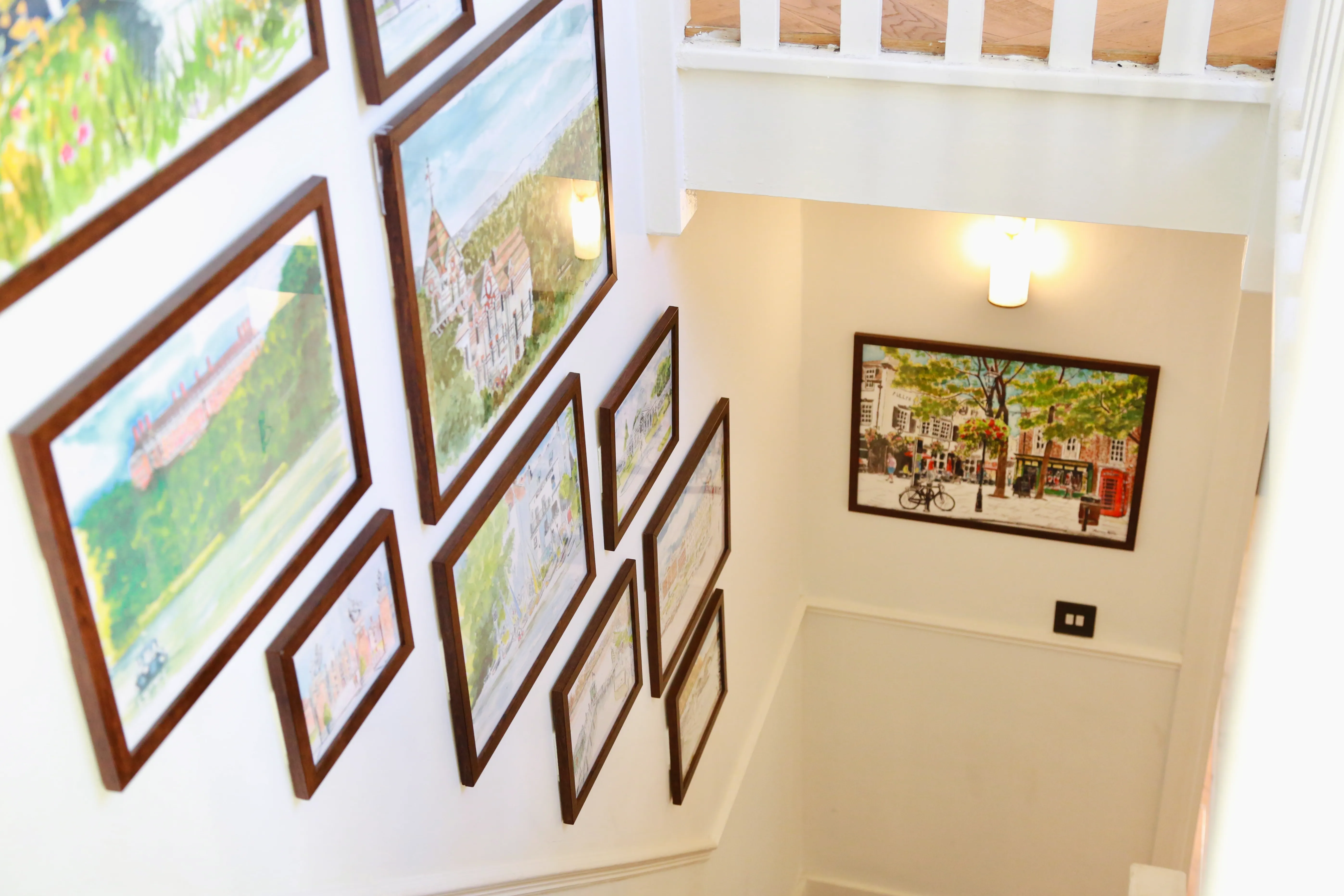 Wall of framed paintings depicting buildings and nature in a stairwell with white walls.