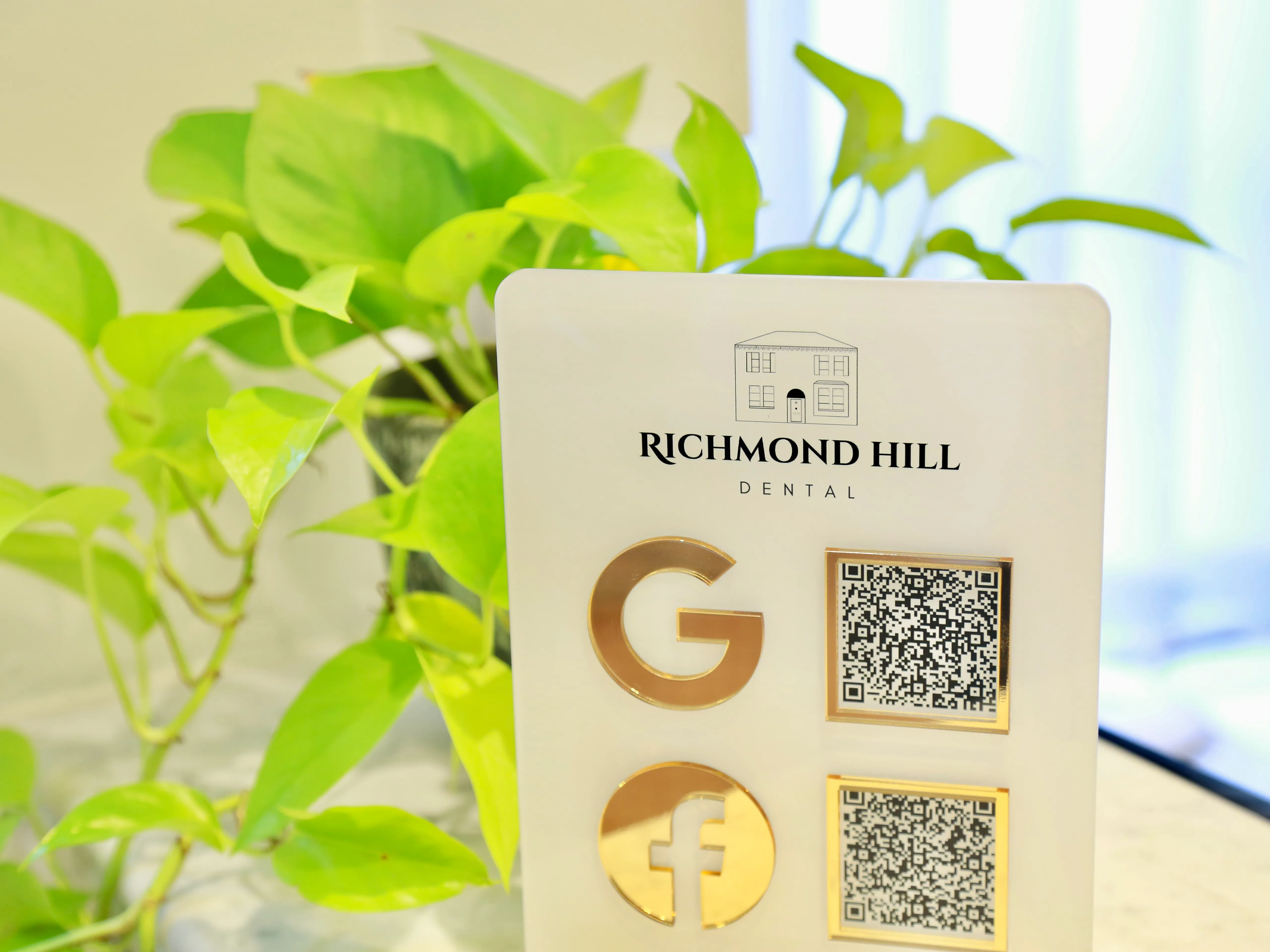Richmond Hill Dental sign with gold Google and Facebook icons and two QR codes, green plant in background.