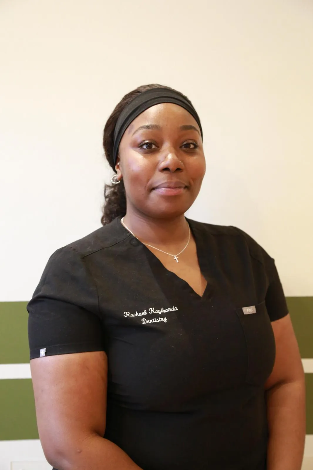 Woman in black dentistry scrubs and headband standing against a light wall with green stripe.