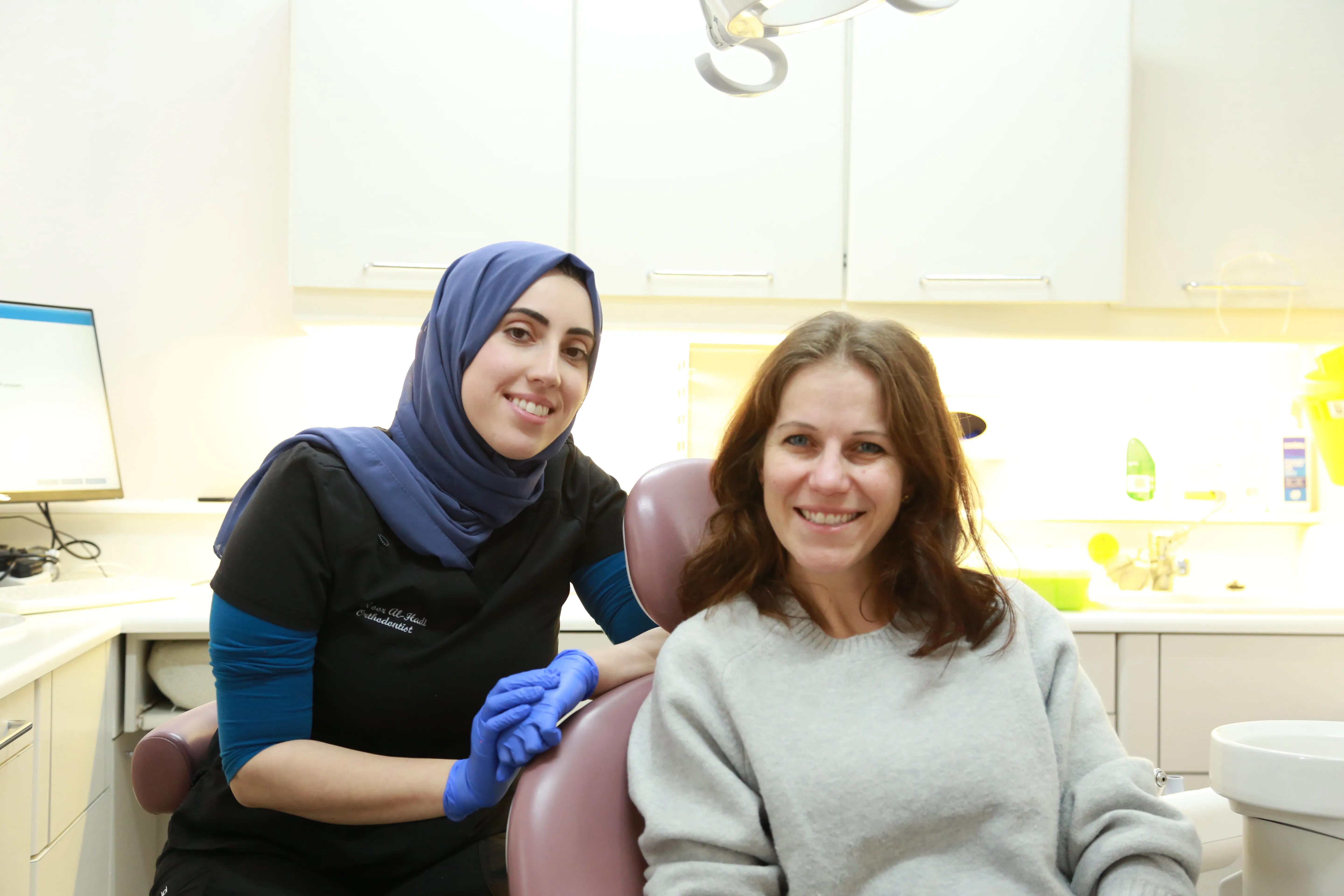 Smiling female dentist in hijab with blue gloves stands next to a seated female patient in dental office.