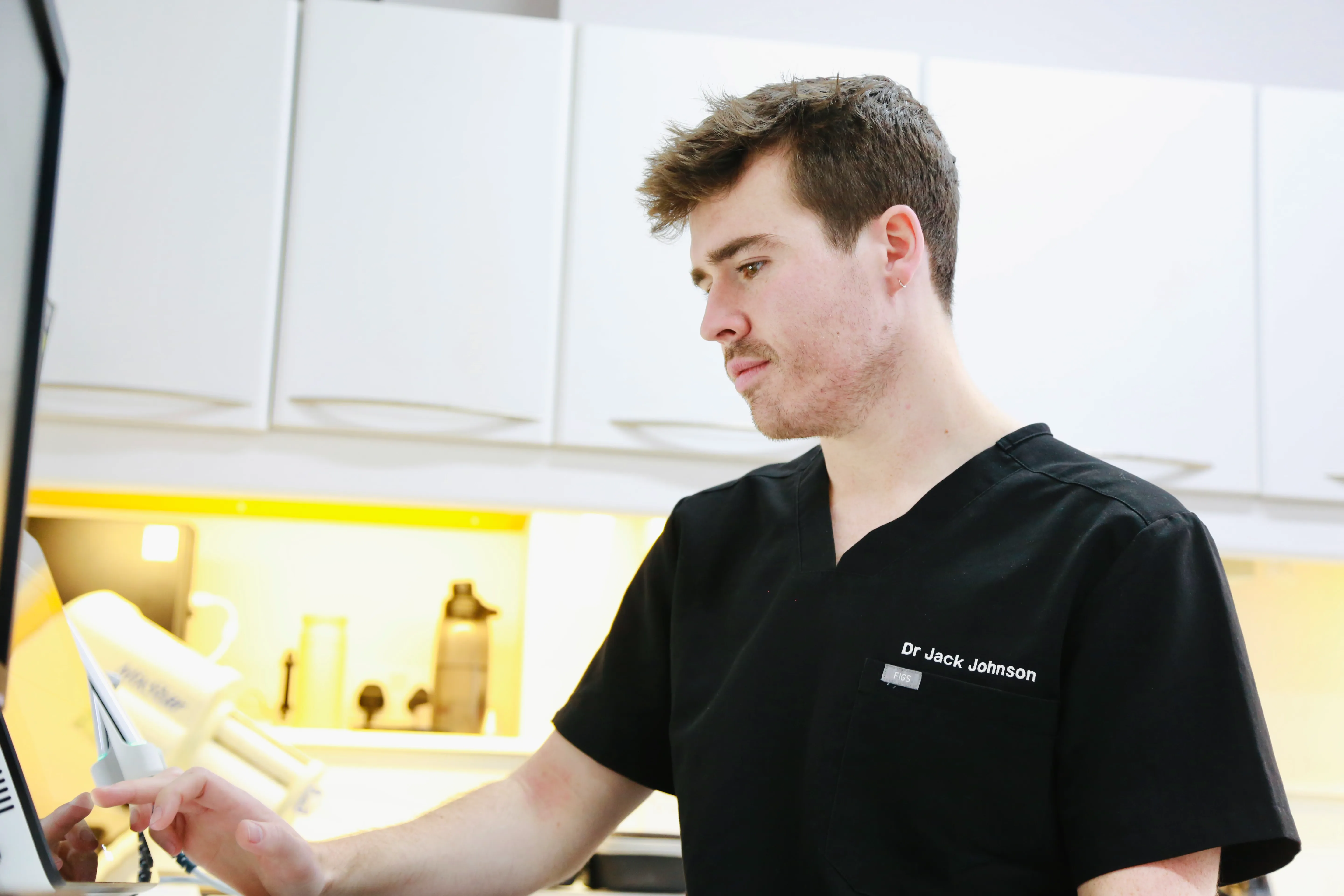 Male doctor in black scrubs named Dr Jack Johnson using a touchscreen monitor in a medical office.