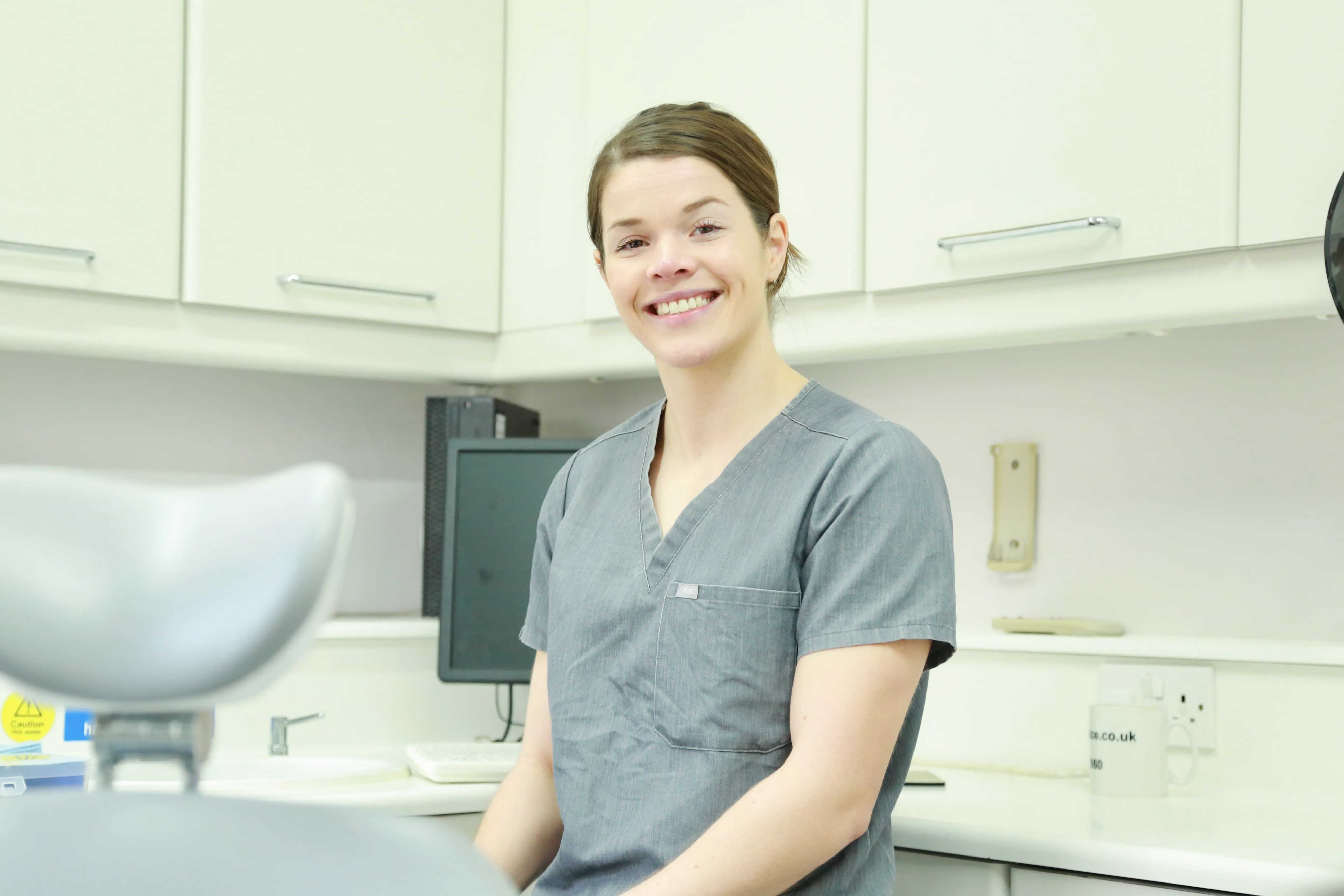 Smiling female medical professional in gray scrubs seated in a clean, white clinical room.