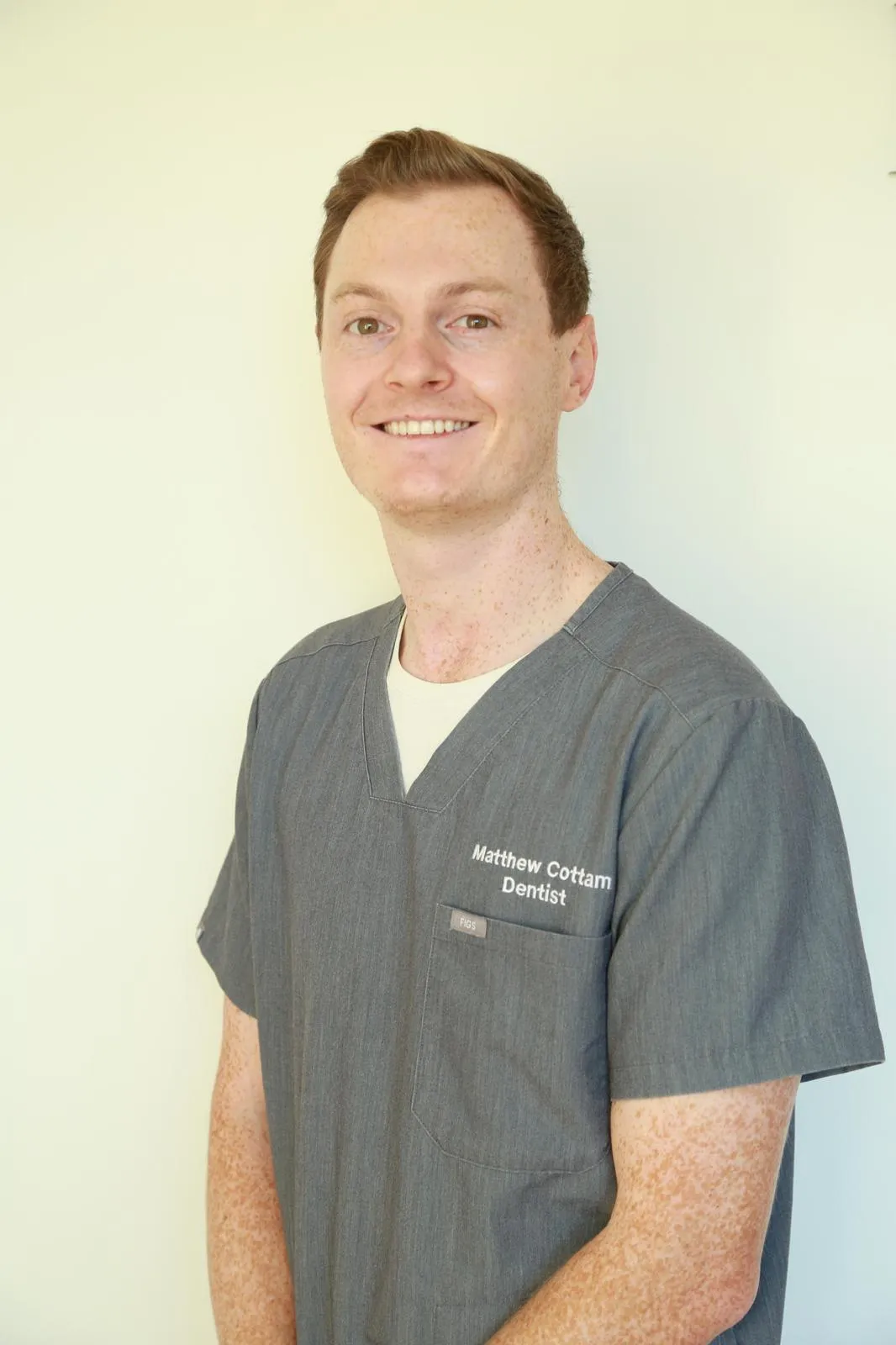 Smiling man with fair skin and freckles wearing grey scrubs labeled Matthew Cottam Dentist.