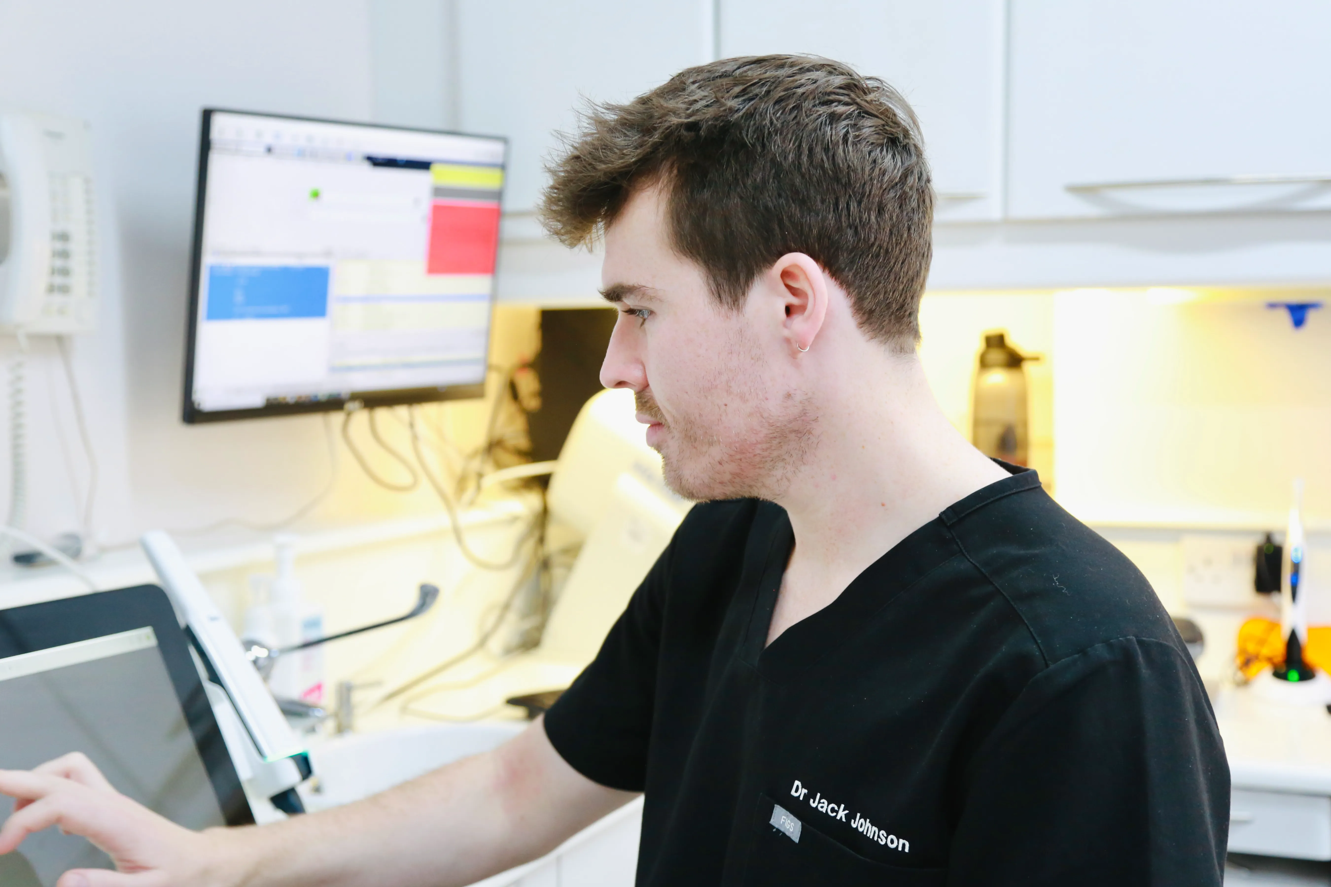 Young male doctor in black scrubs using a touchscreen device in a clinical room.