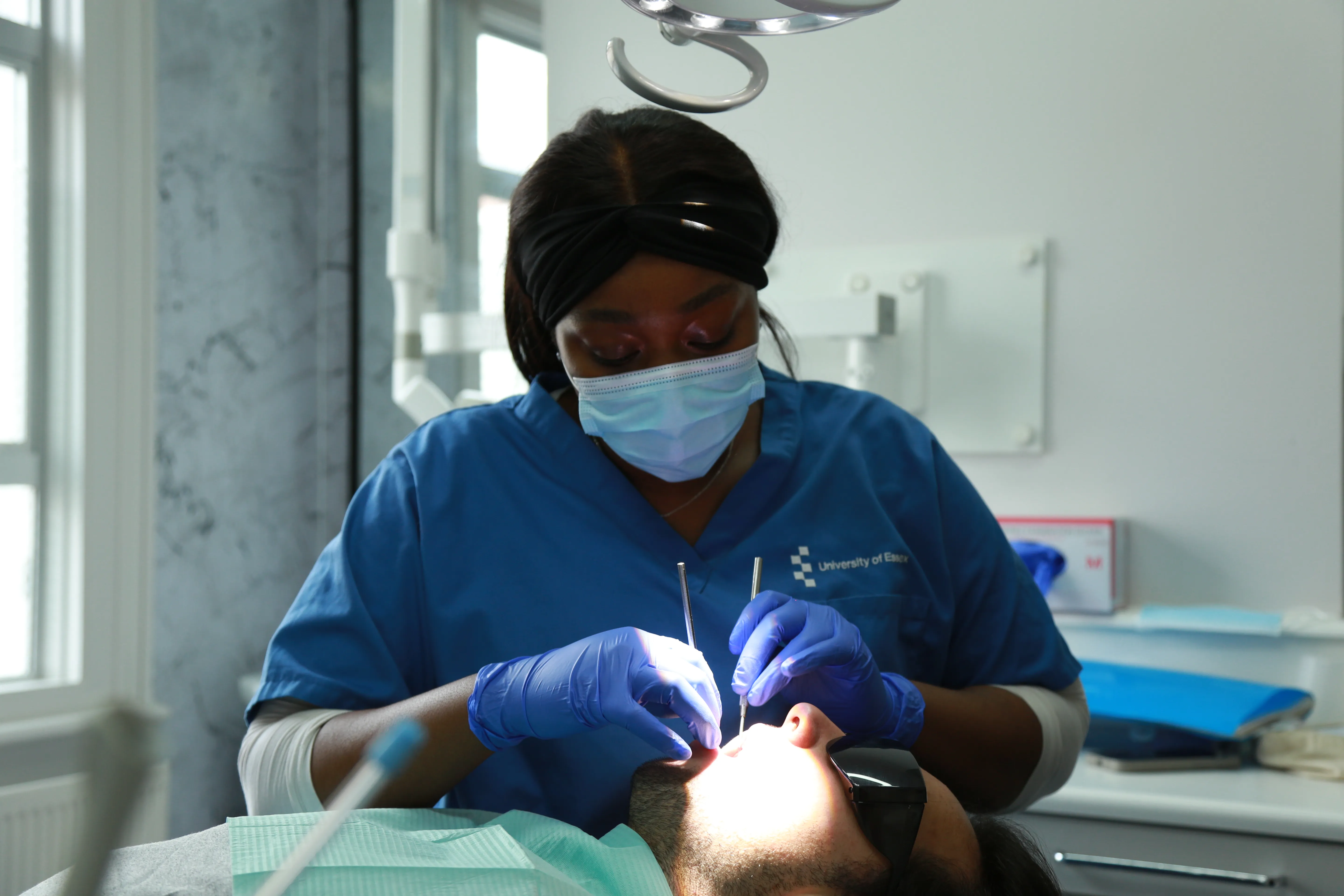 Dentist wearing blue scrubs and mask performs dental exam on a male patient lying back in chair.