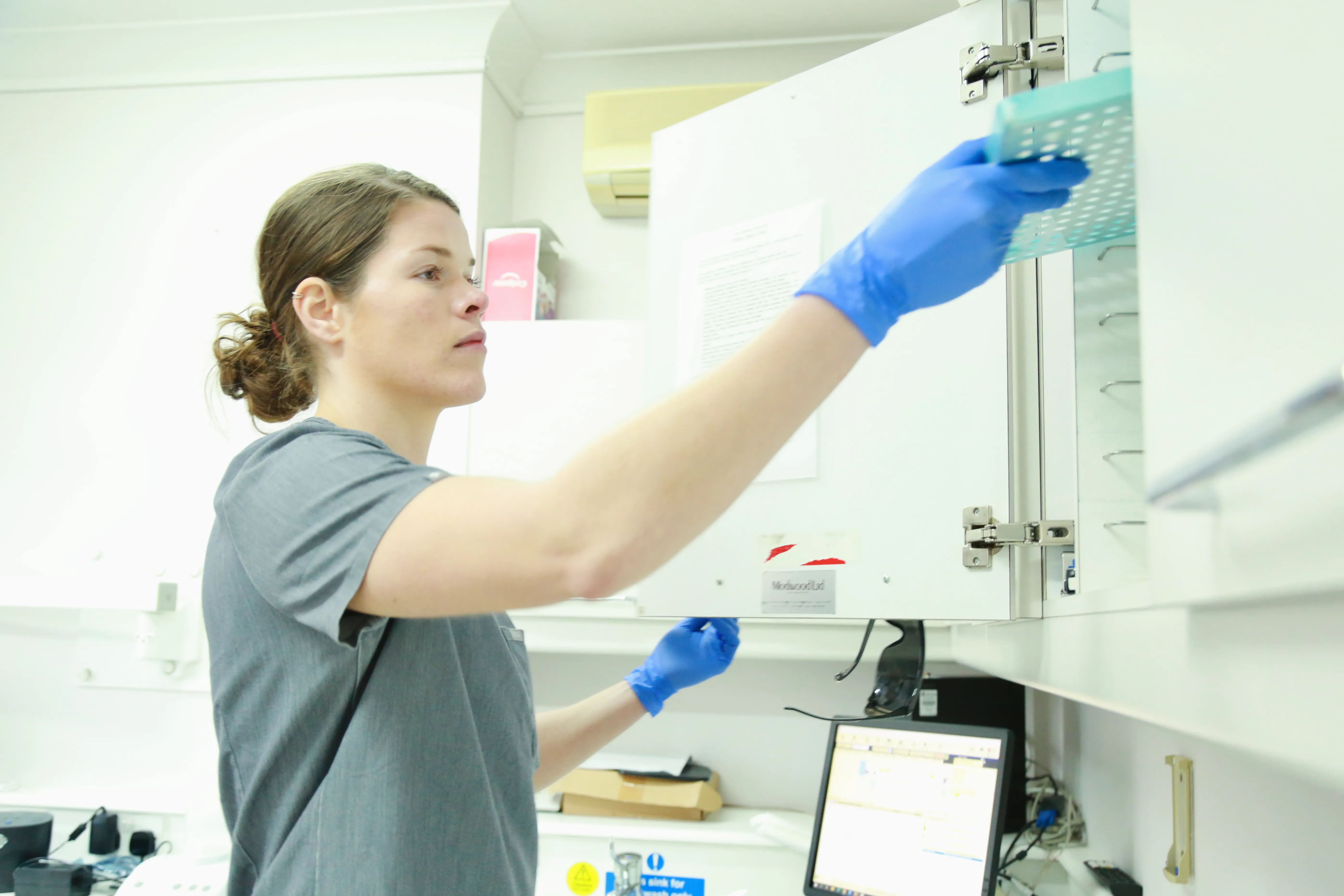 Person wearing blue gloves and gray scrubs placing a tray in a white cabinet in a lab.
