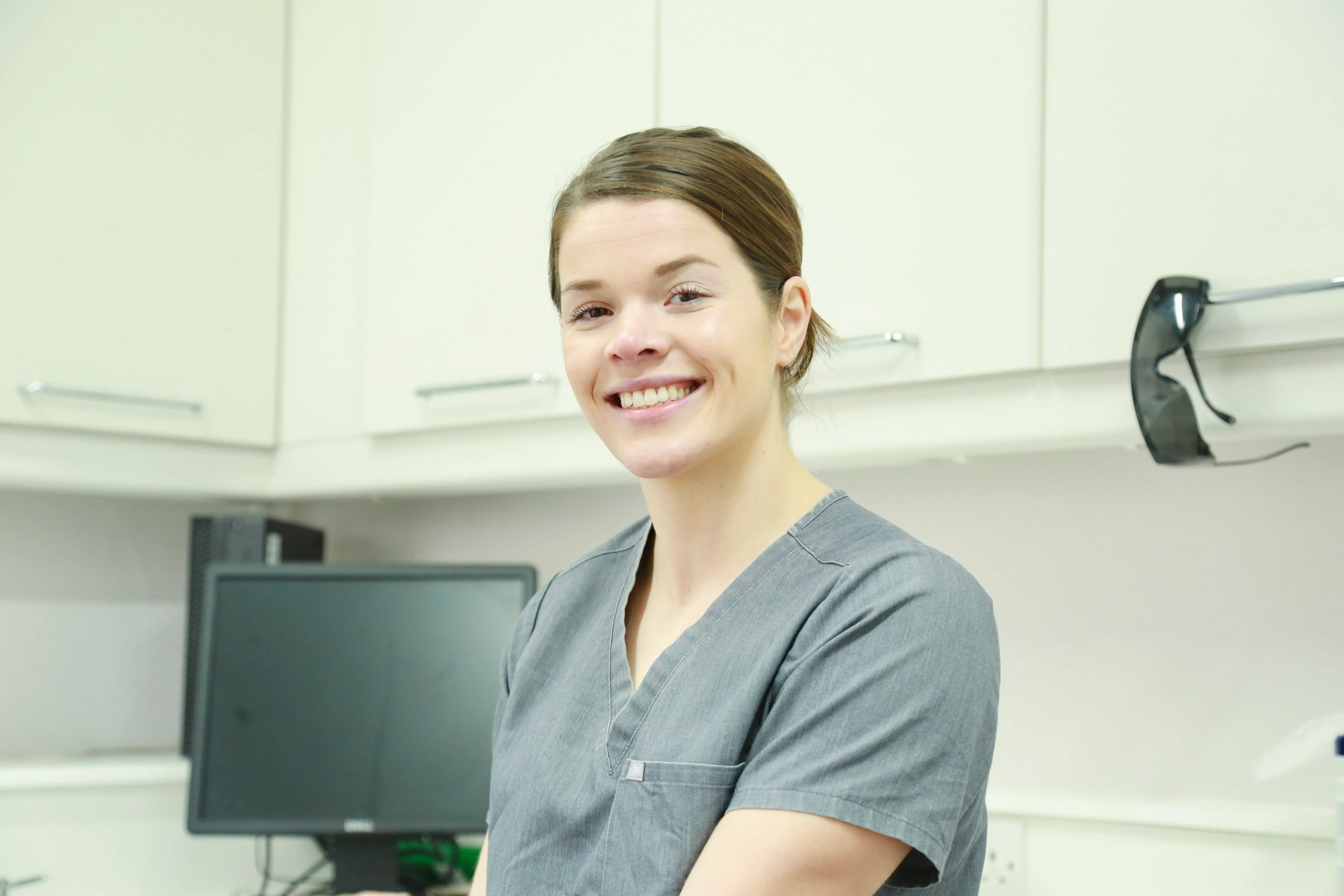 Smiling female nurse or medical professional wearing gray scrubs in a clinical setting.