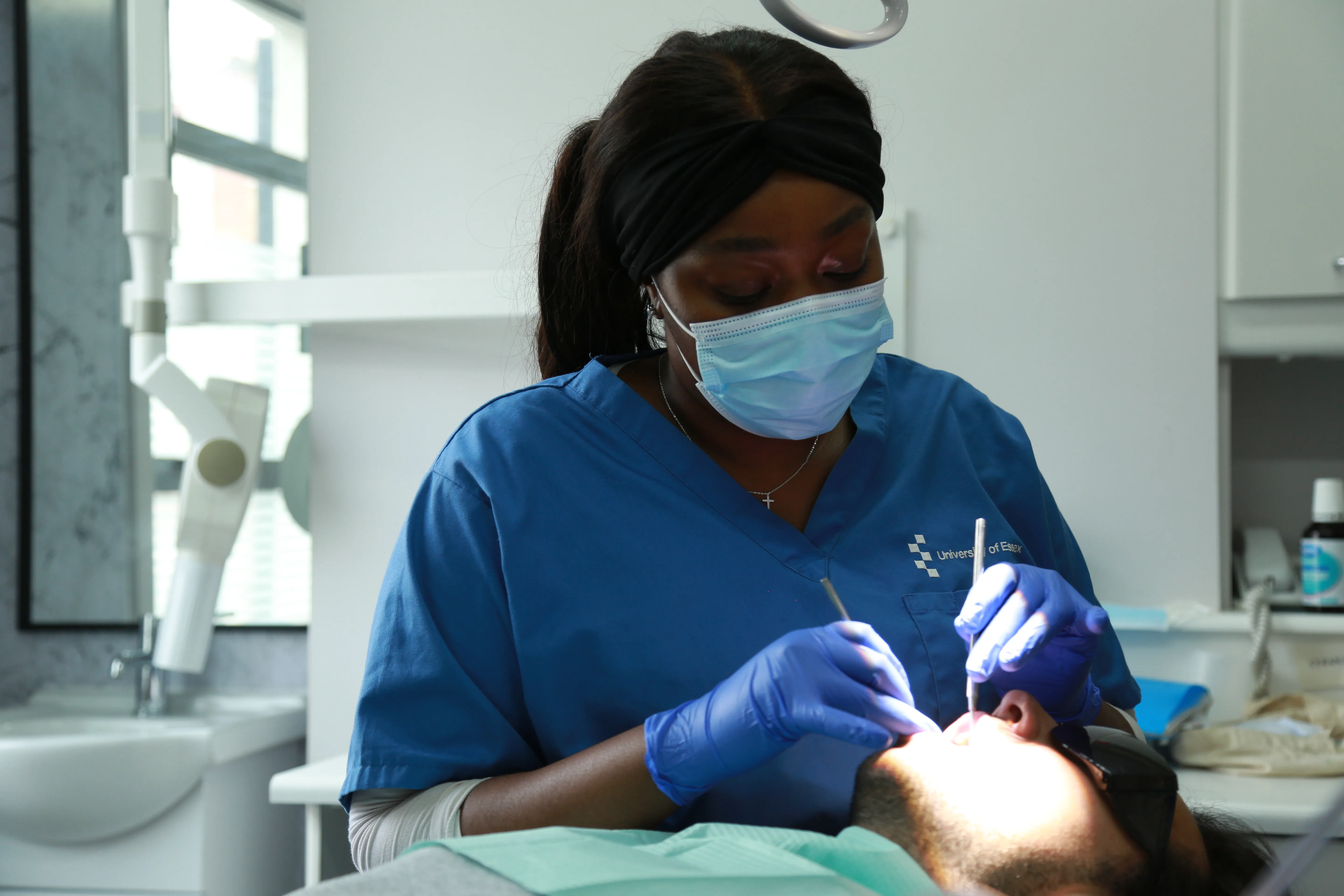 Dentist in blue scrubs and mask examining a patient's mouth under bright light in a clinic.