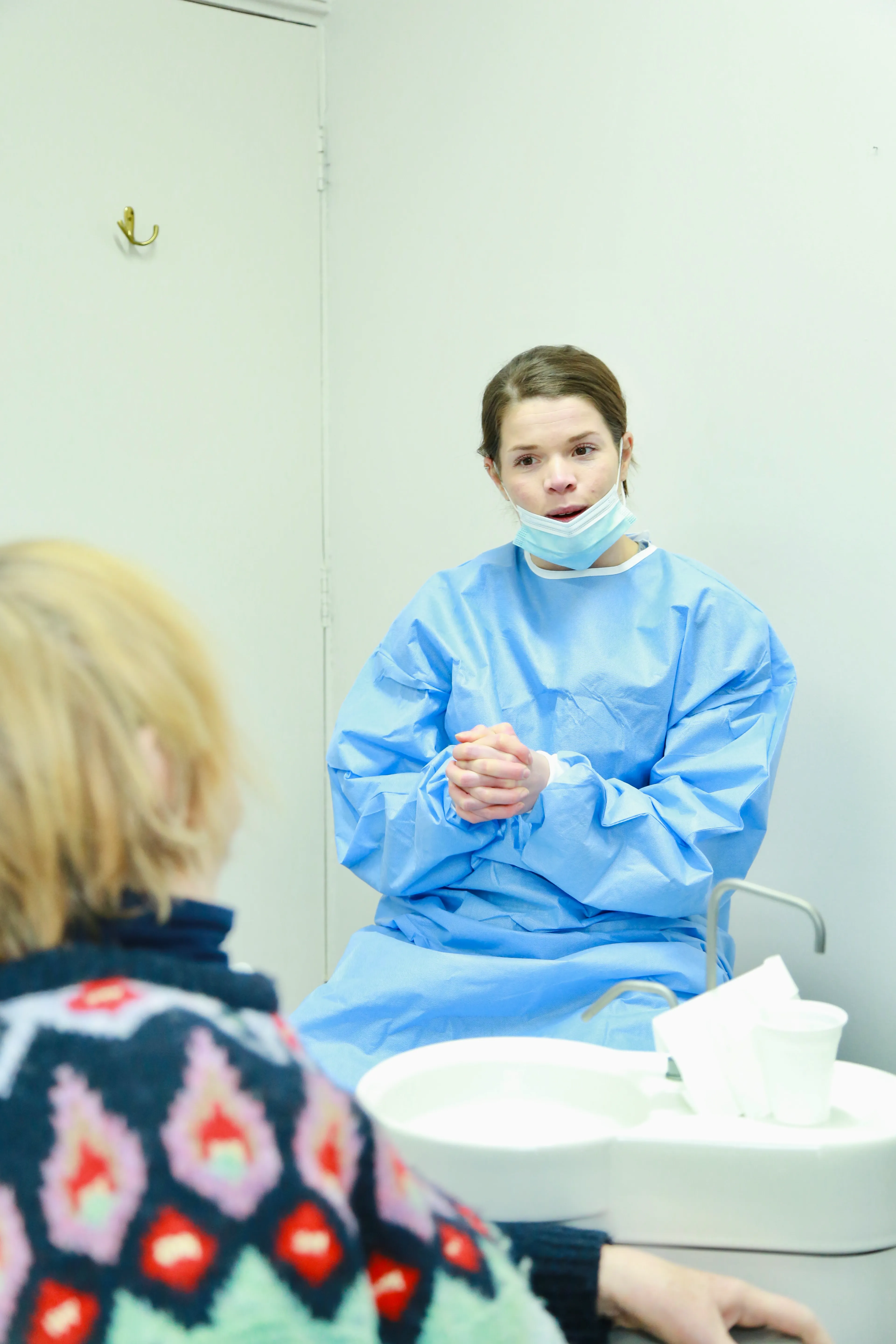 Healthcare worker in blue gown and mask talks to patient in colorful sweater in clinical room.