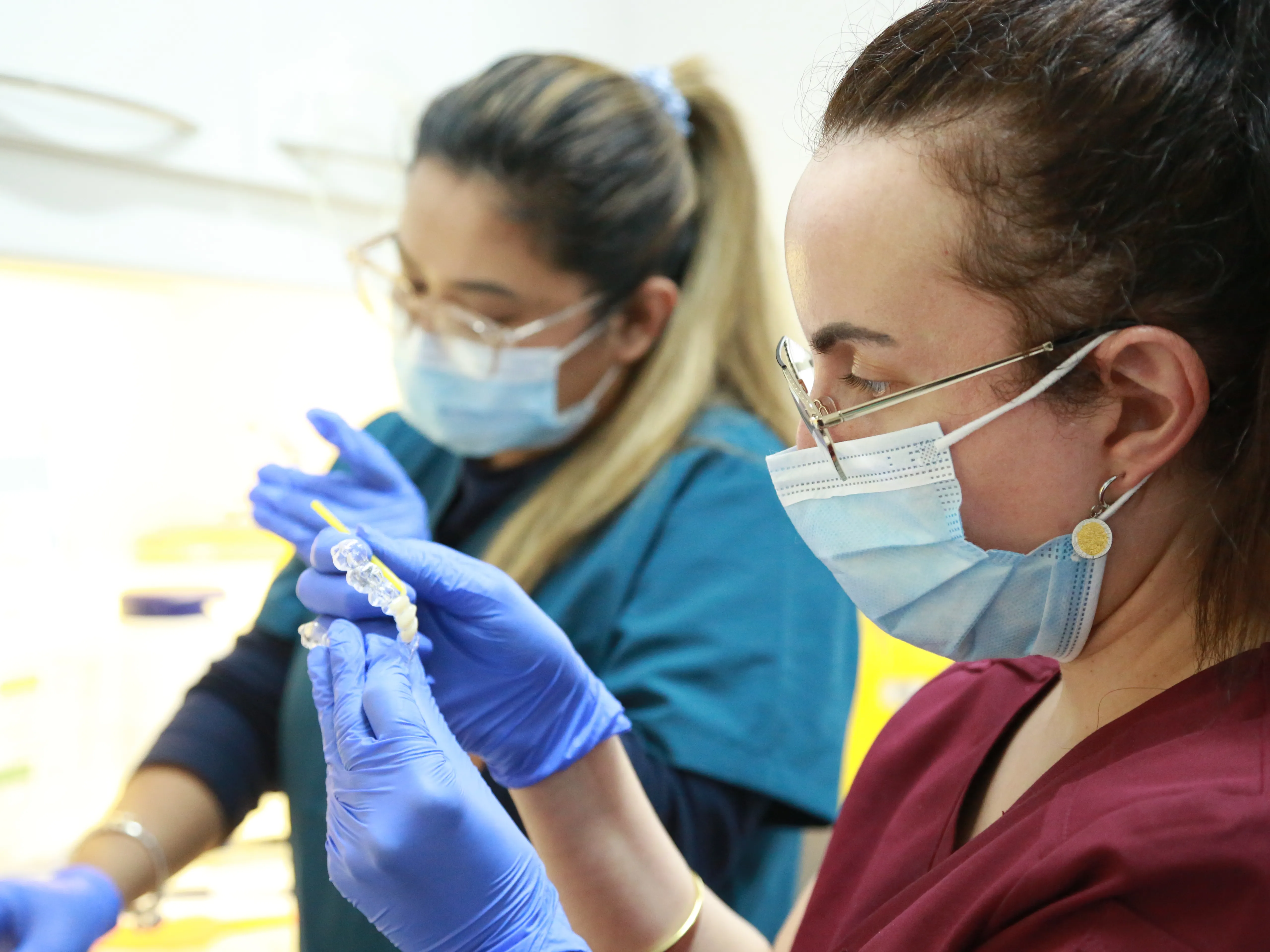 Two dental professionals wearing masks and gloves examine clear dental molds closely.
