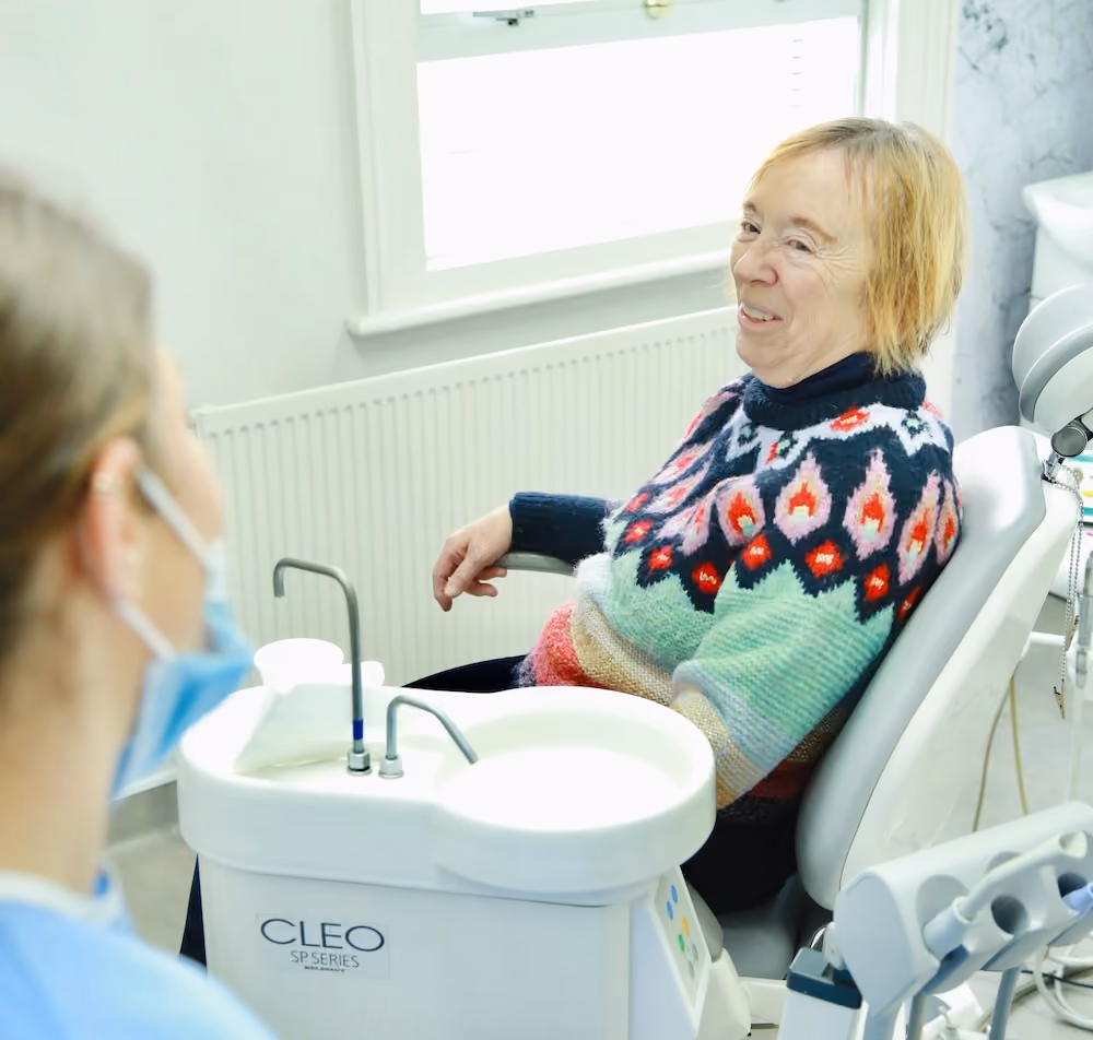 Older woman smiling while seated in a dental chair with a dental professional nearby wearing a mask.