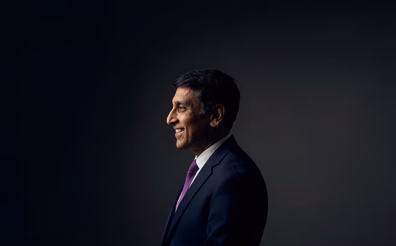 Profile portrait of a smiling man in a navy suit and purple tie against a dark background