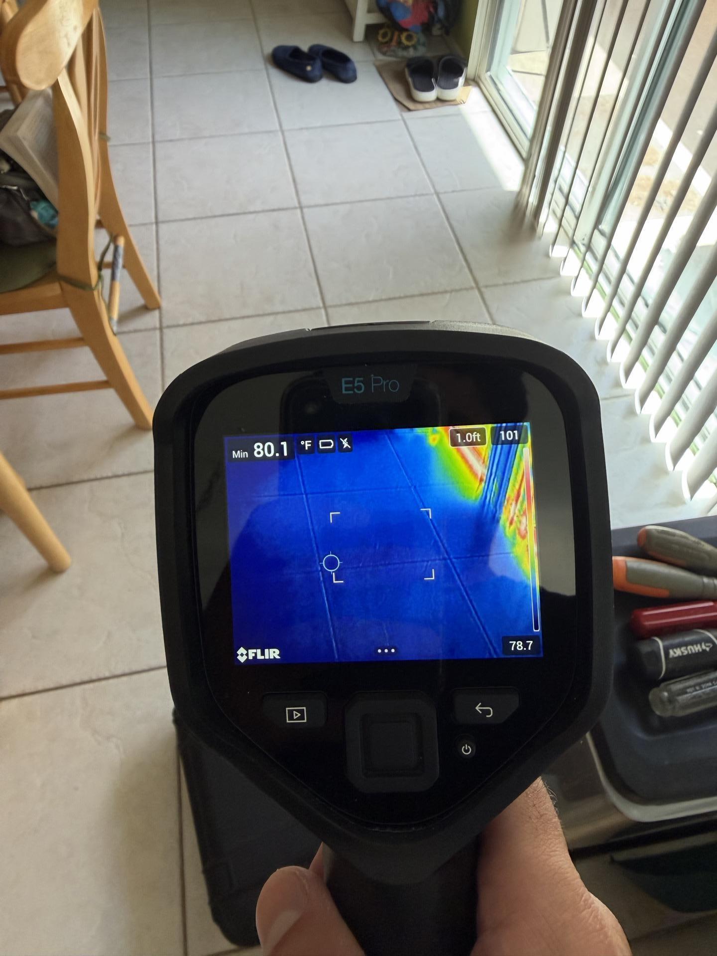 A thermal camera held over a tiled floor near a sliding glass door, its screen showing a blue and yellow heat scan.