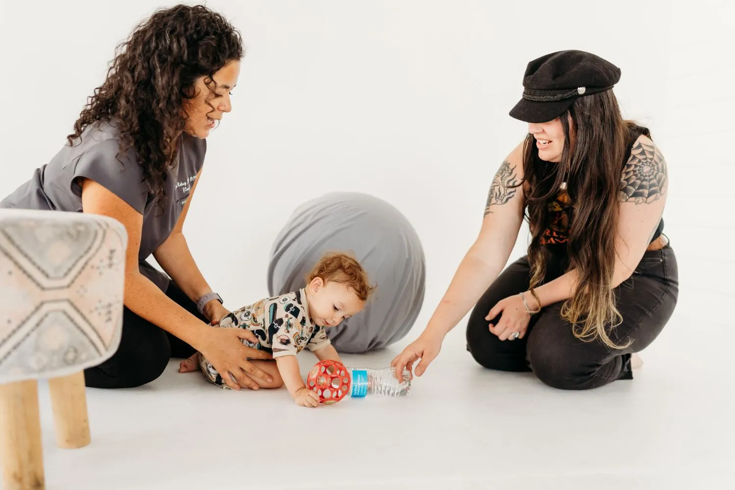 Two women on floor with a toddler playing with a red ball and a plastic water bottle.