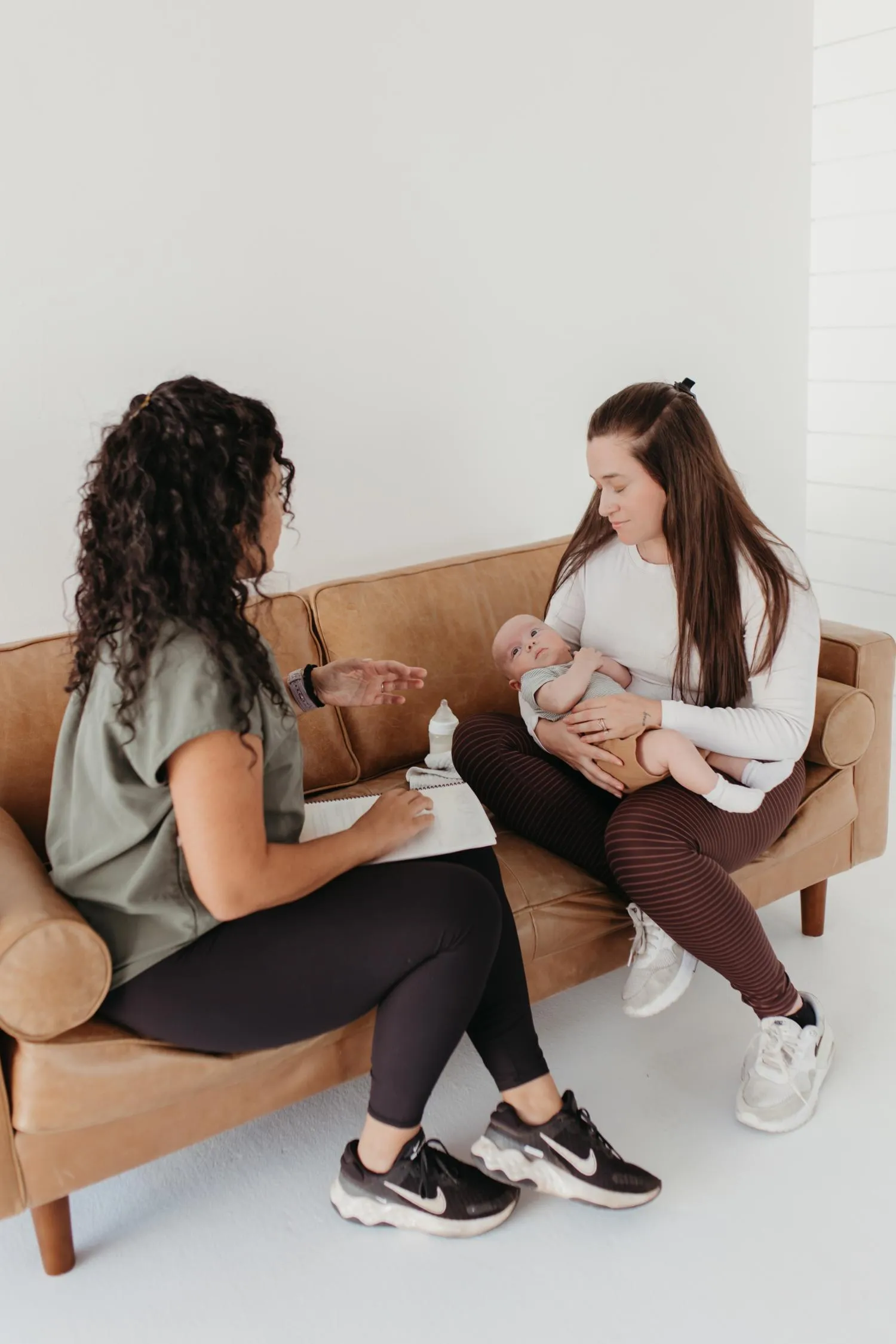 Woman sitting on a couch holding a baby talks with another woman taking notes.