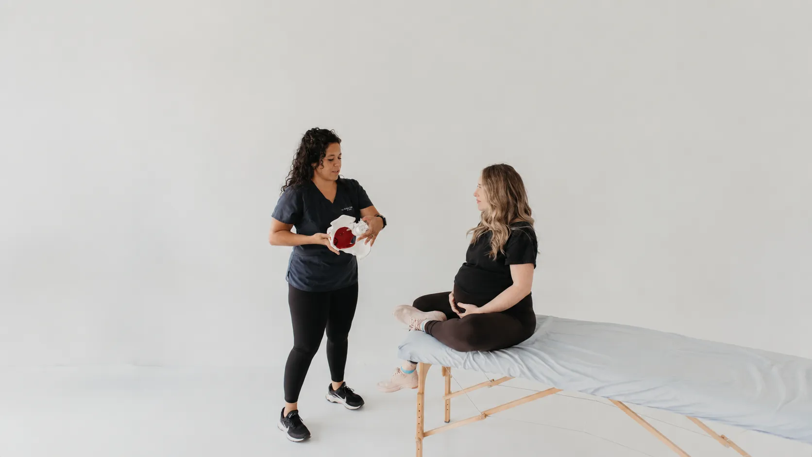 Pregnant woman sitting on an exam table listening to a healthcare professional holding a pelvic model.