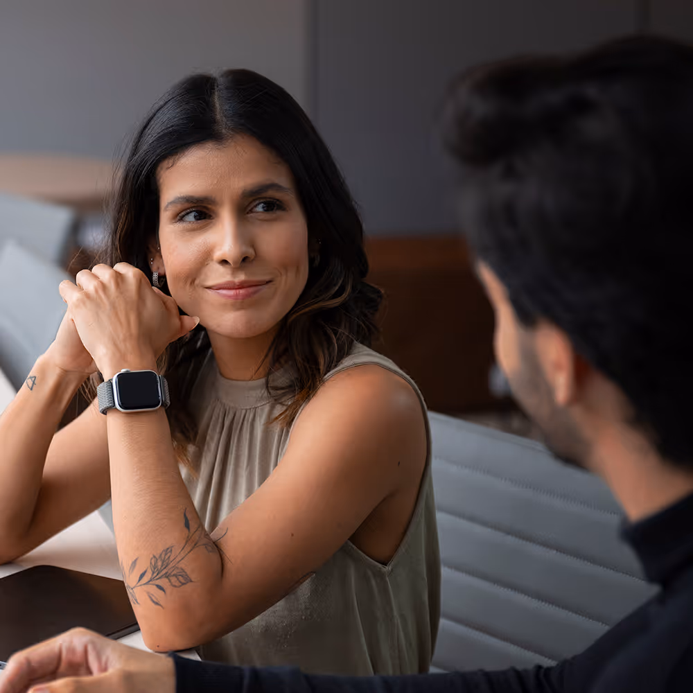 Woman with floral tattoo and smartwatch smiling, sitting and talking to a man at a table.