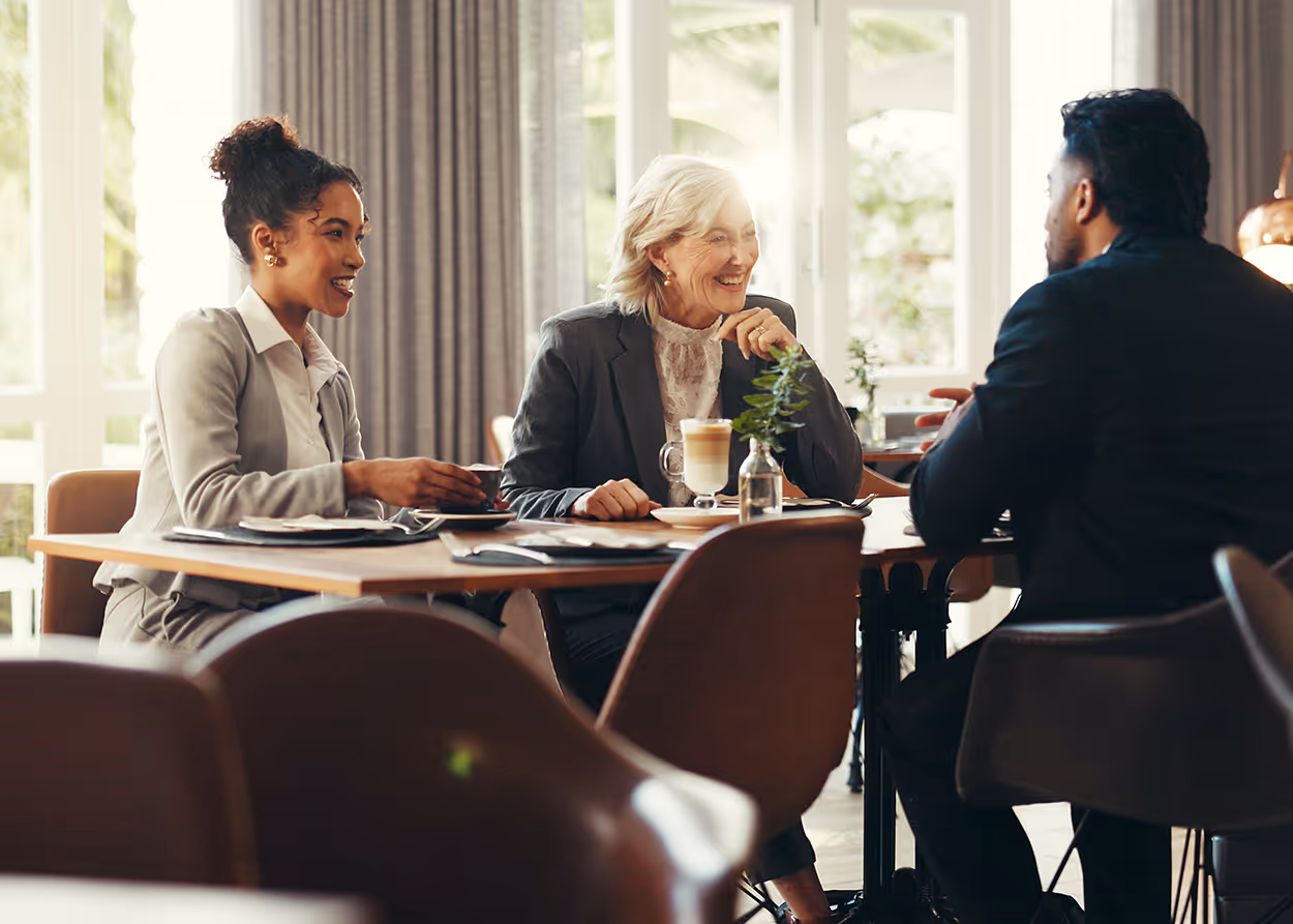 Three business people in professional attire chatting and drinking coffee at a cafe table during daylight.