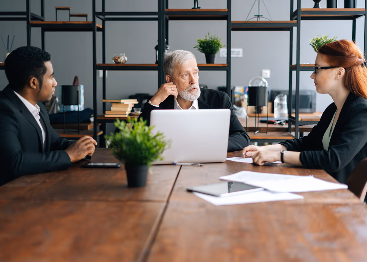 Three businesspeople in a meeting around a table with a laptop, papers, and a potted plant.