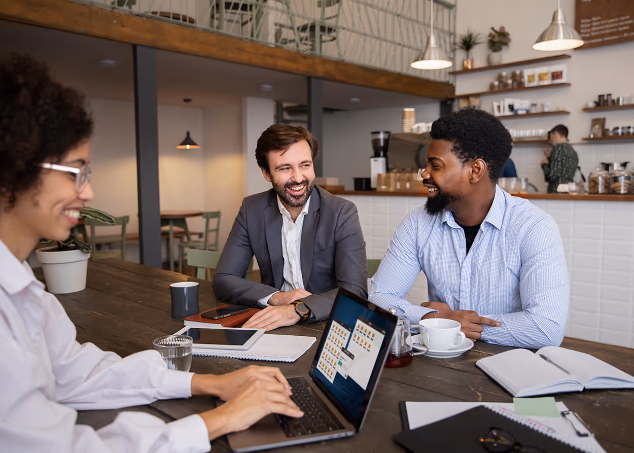 Three professionals having a meeting at a table with laptops, notebooks, and coffee cups in a cafe.